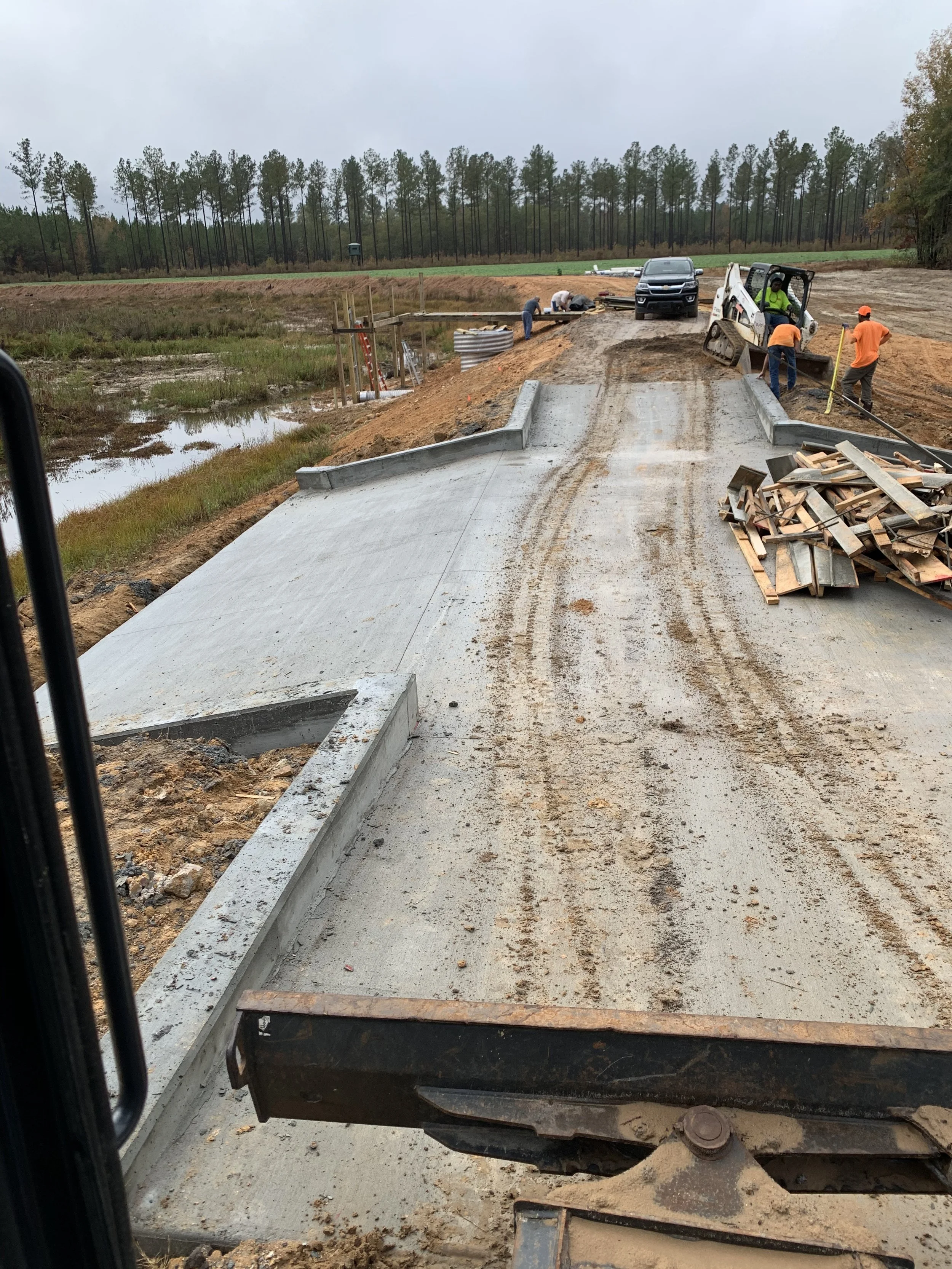 Construction workers working on a new bridge or road, with equipment and a vehicle nearby, in a rural area with trees and marshland in the background.