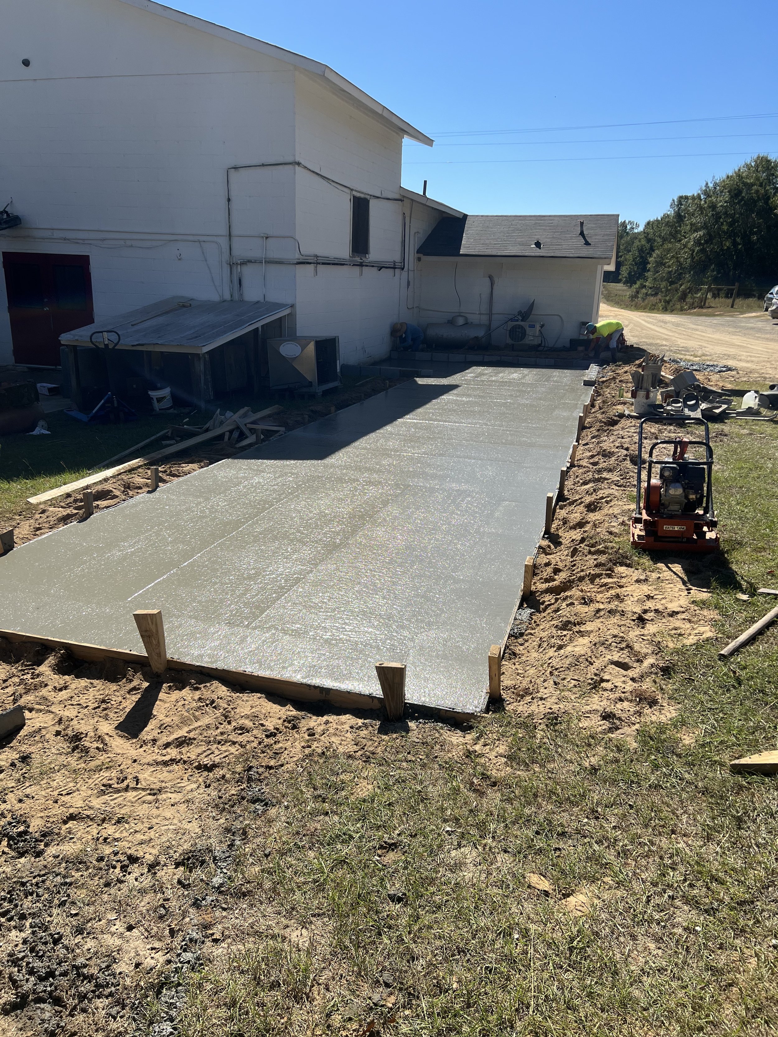 Concrete slab being poured for a new outdoor patio or walkway, construction site with tools and equipment, house in the background.