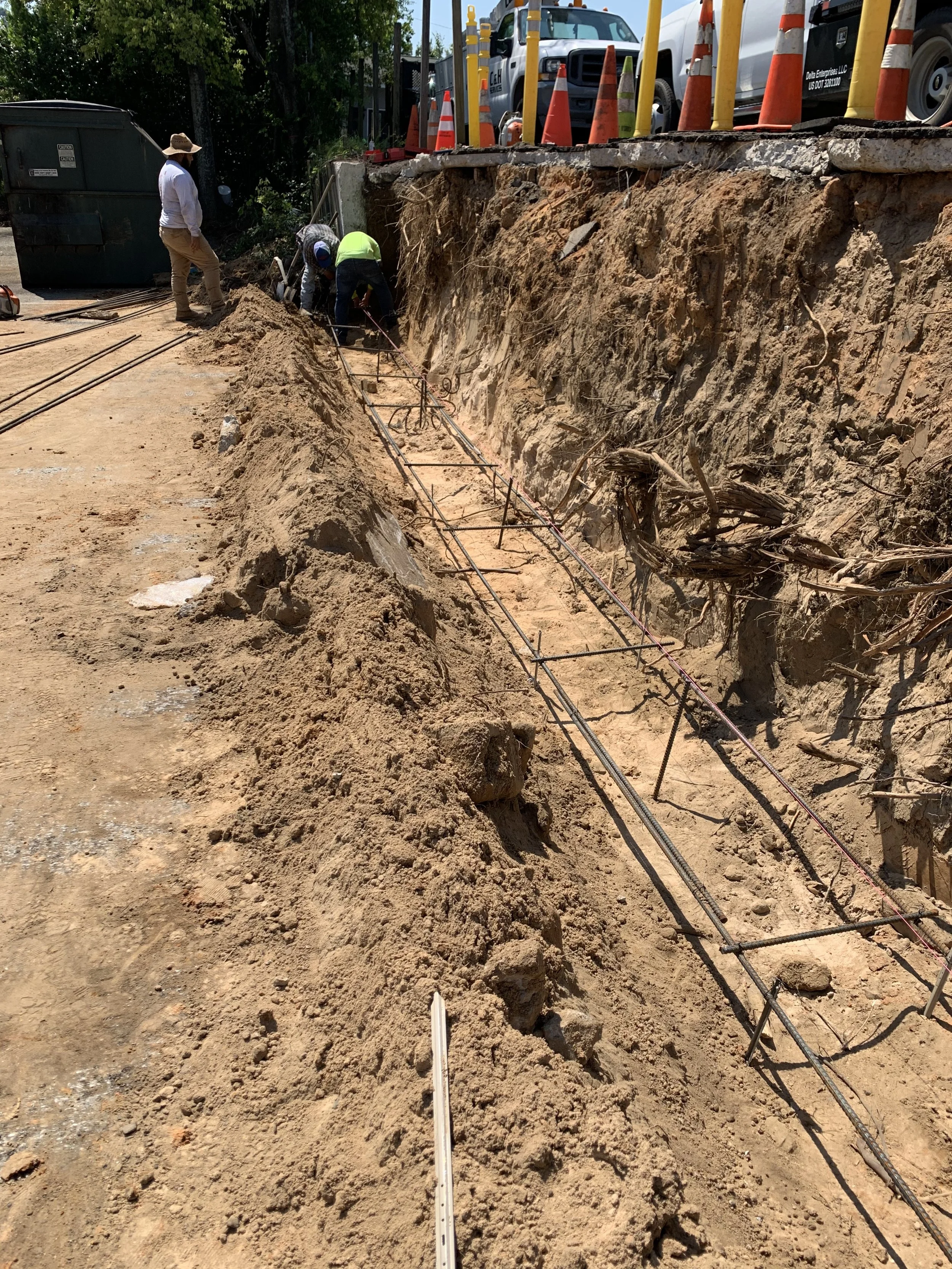 Construction workers installing rebar in a trench on a sunny day, with orange safety cones and trucks nearby.