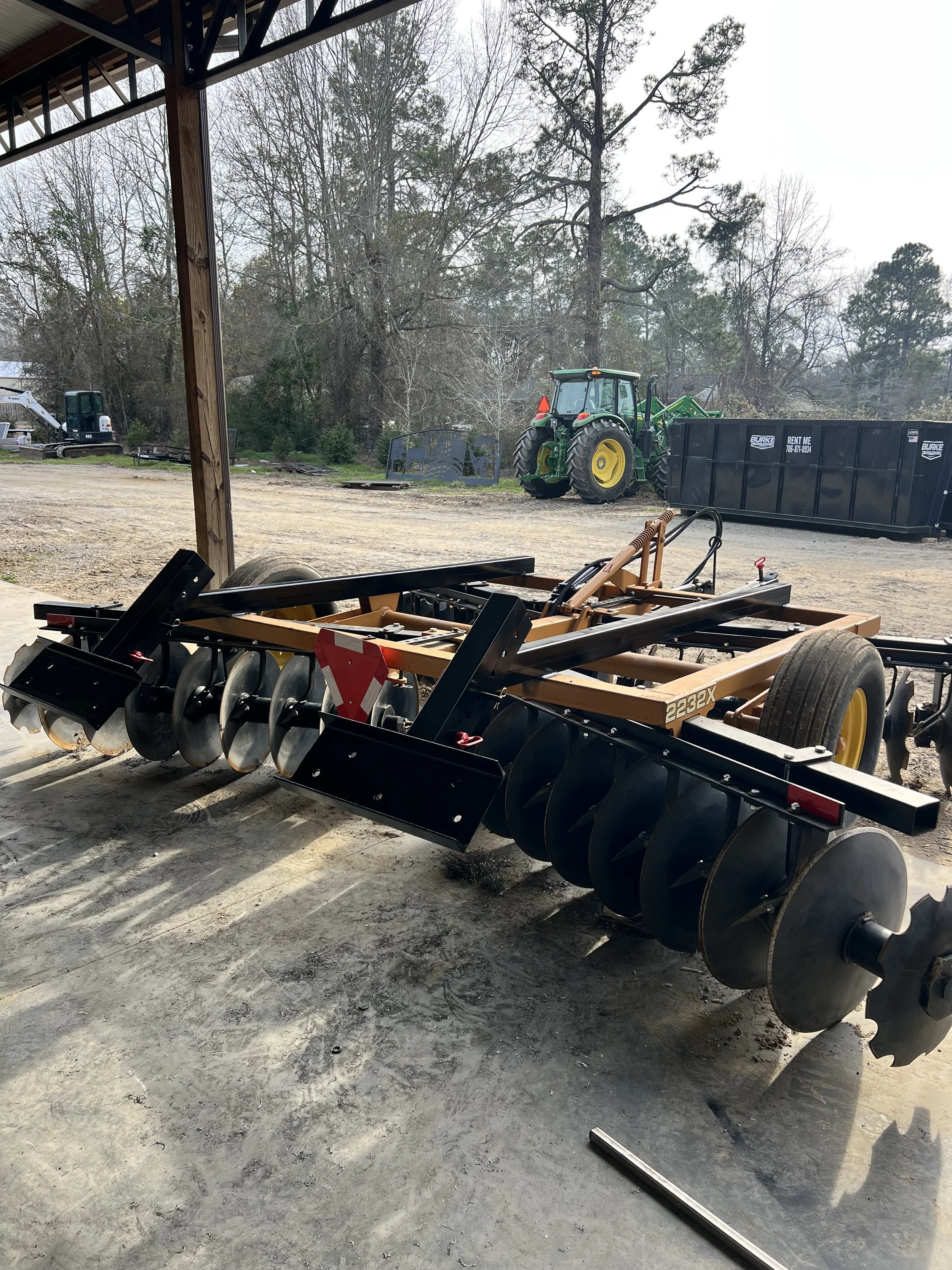 A black and orange disc harrow on a concrete floor under a shed with a green tractor and a black dumpster outside.