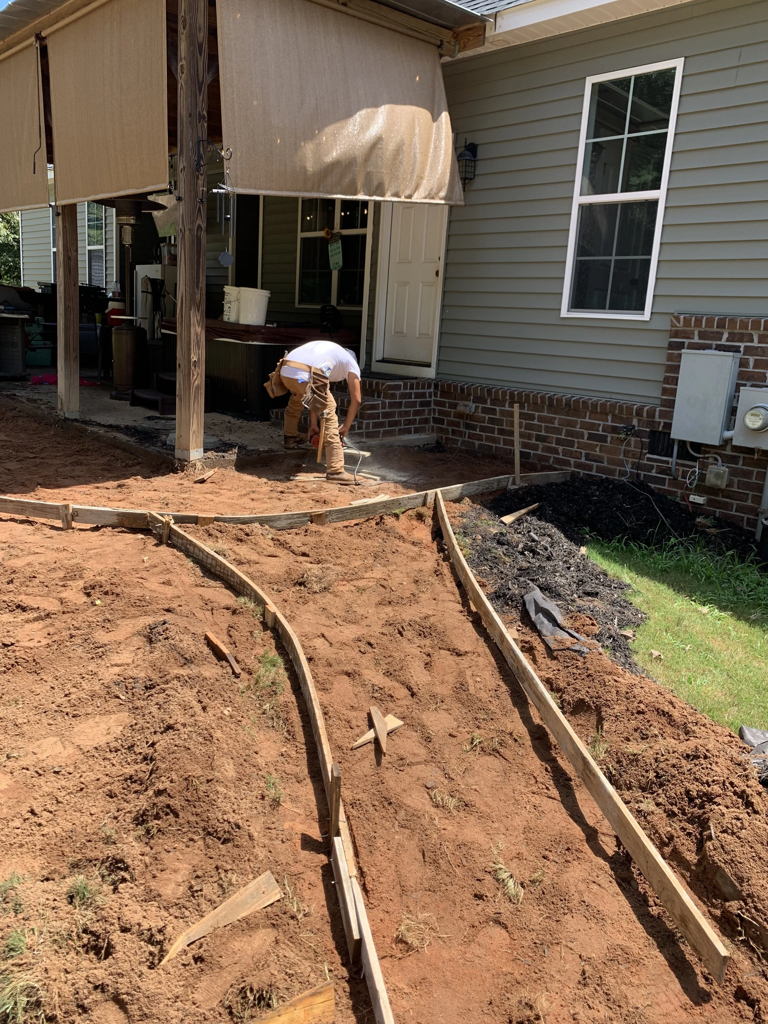 Man working on a backyard landscaping project, laying out wooden boundary borders on reddish soil in front of a house with beige siding and brick foundation, during daytime.