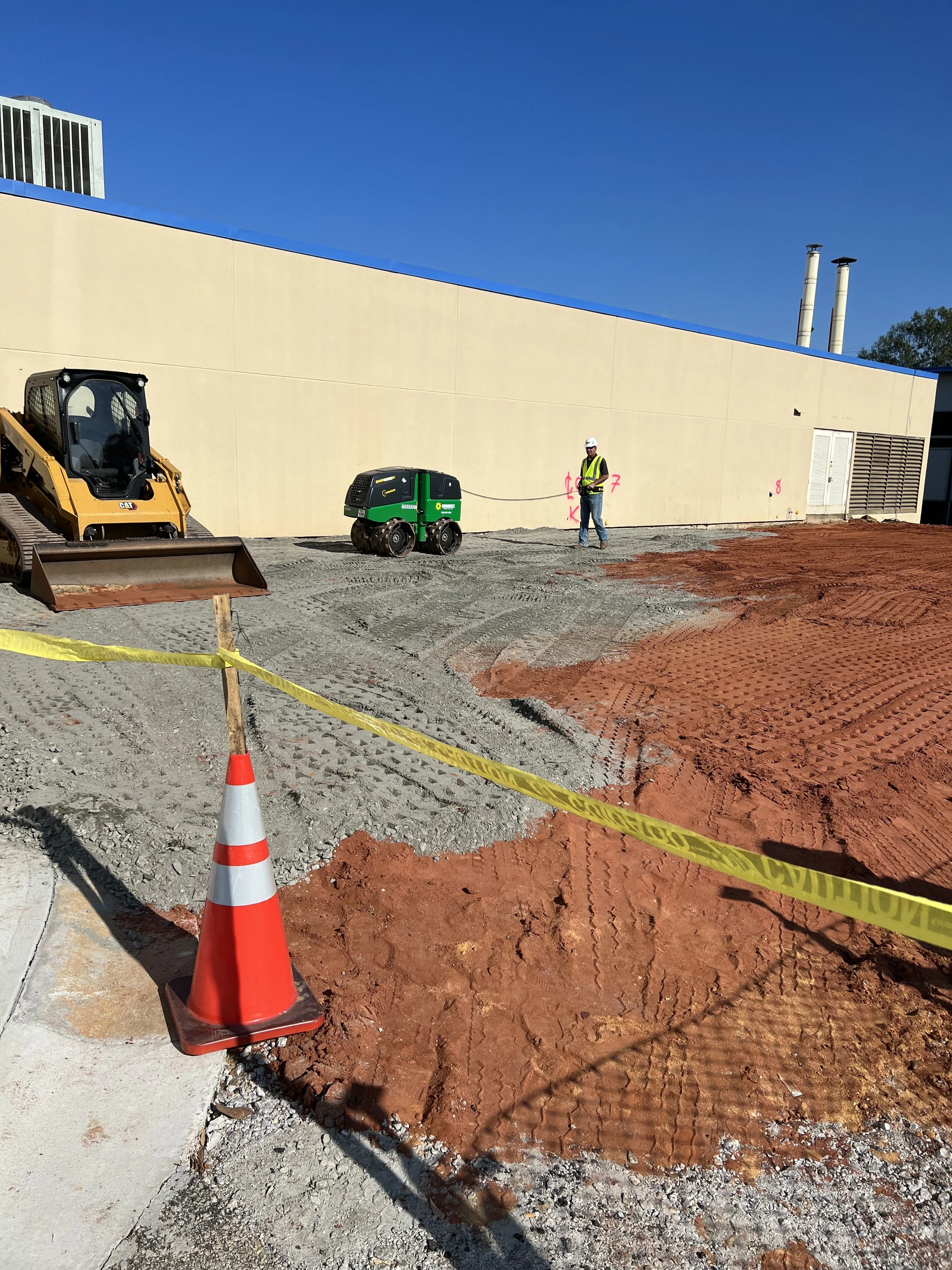Construction site with a worker in a safety vest and hard hat, operating a green and black compact equipment, near a beige building with a blue roof. Orange and gravel ground, yellow caution tape, and an orange traffic cone in the foreground.