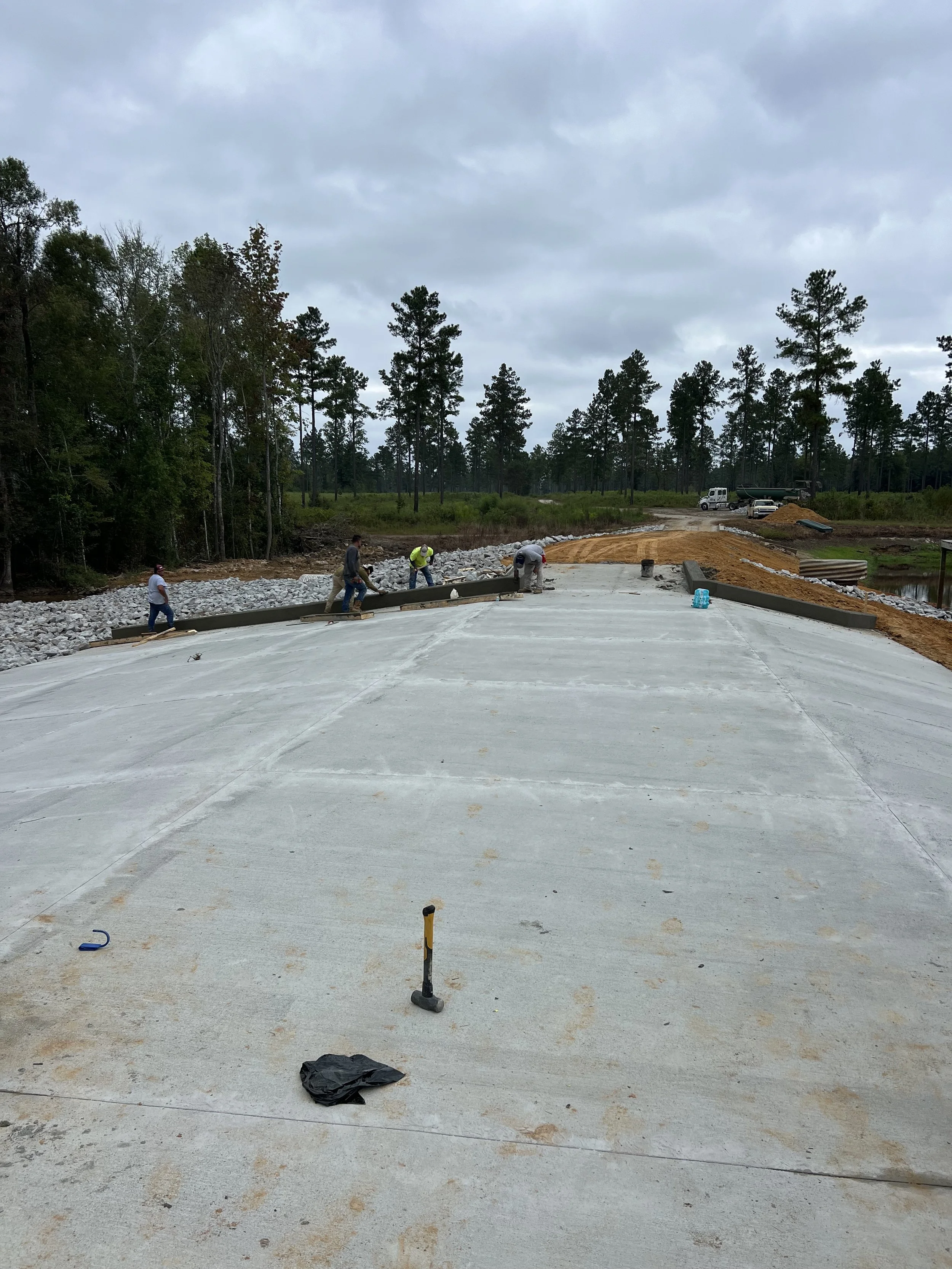 Construction workers are building a road with concrete and gravel, surrounded by trees and a cloudy sky.