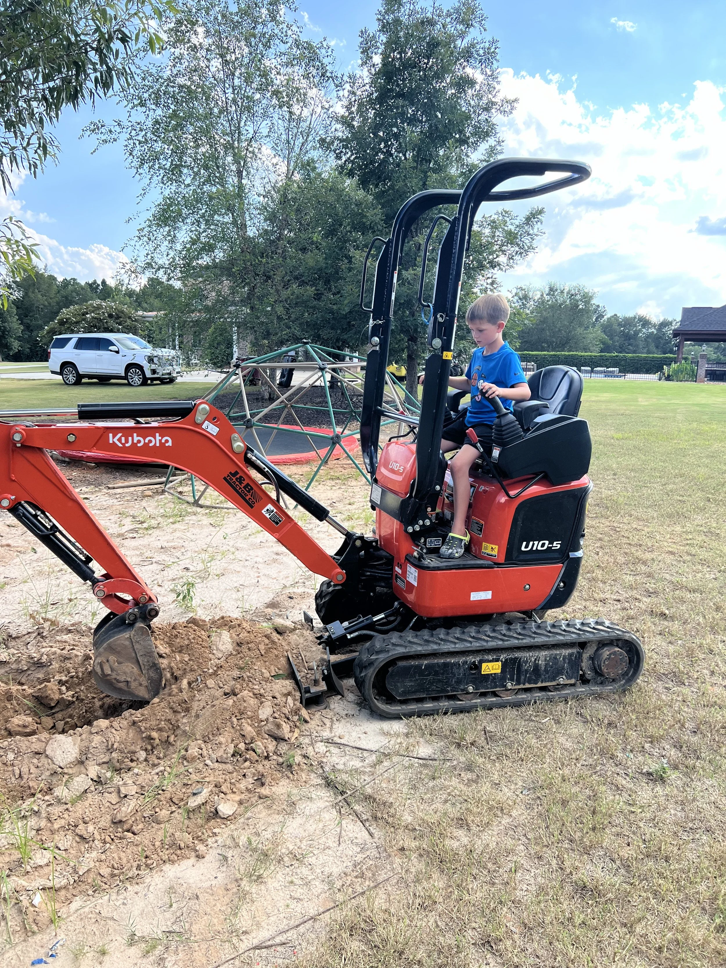 A young boy operating a small Kubota mini excavator on a construction or digging site in a park-like area with trees, grass, a cloudy sky, and a white vehicle in the background.