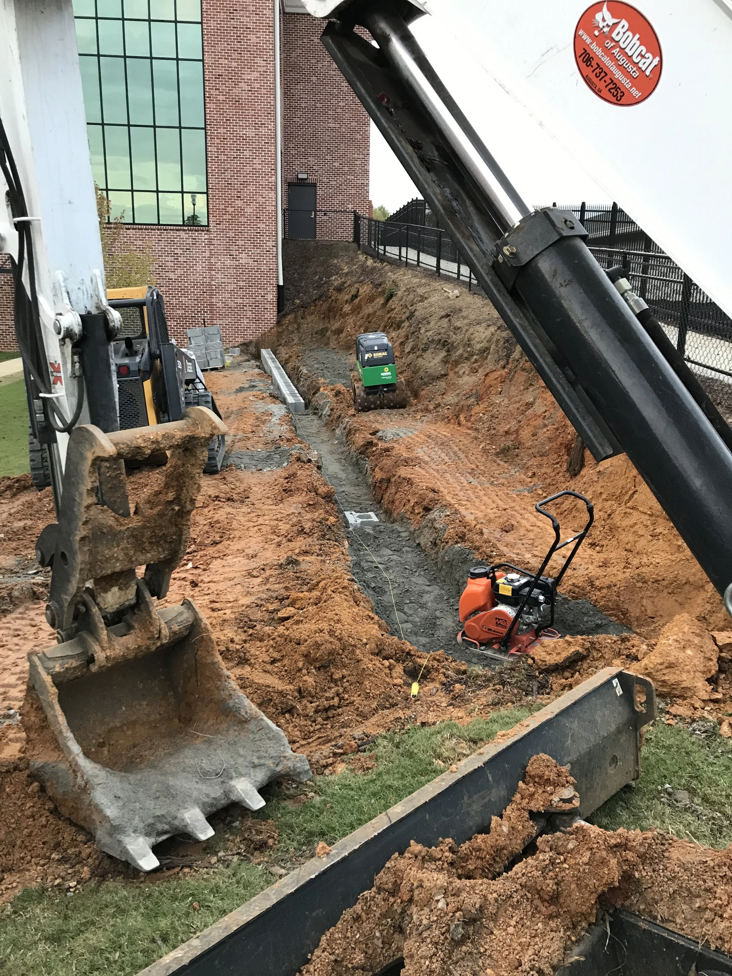 Construction site with excavation equipment, including an excavator bucket, mini-steamroller, and jackhammer, in front of a brick building during daytime.