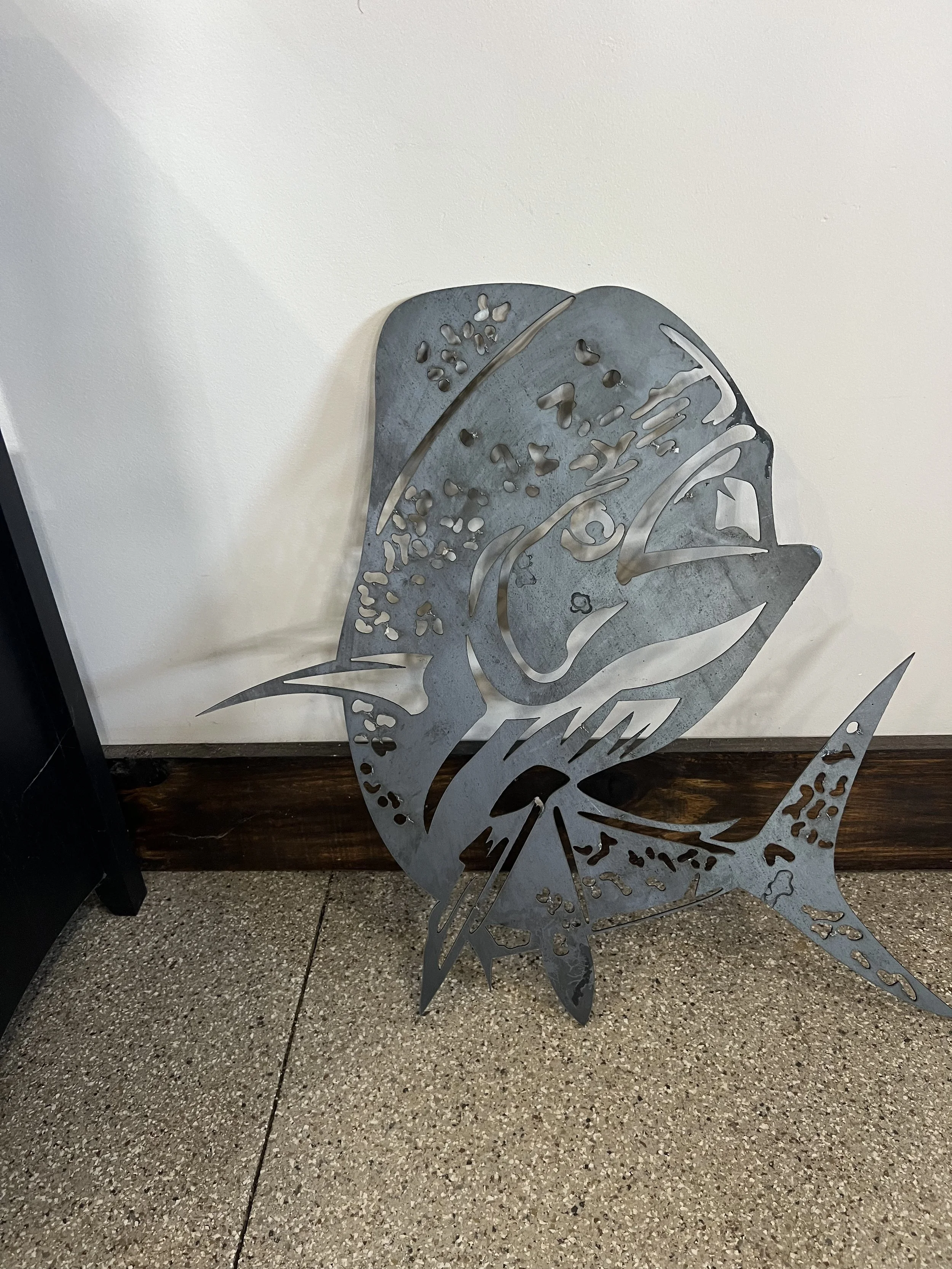 Metal sculpture of a fish, leaning against a white wall on a tiled floor.