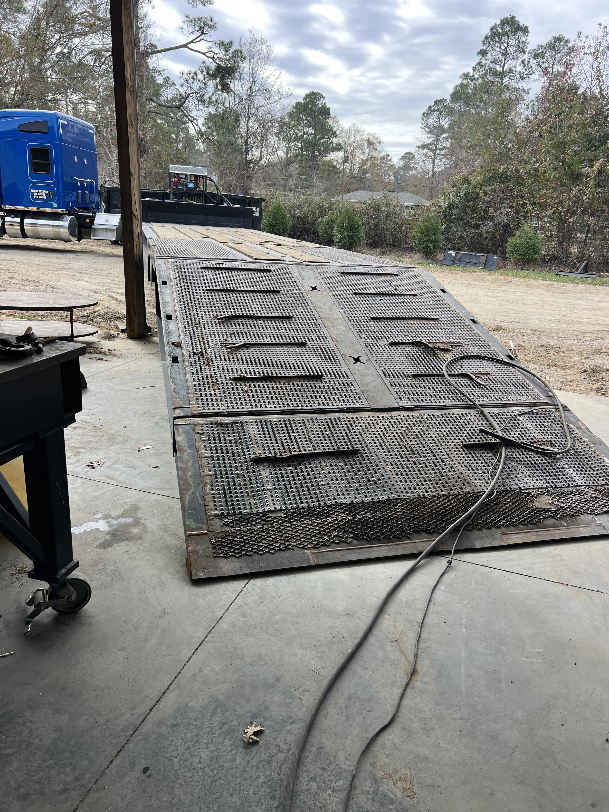A large metal flatbed trailer with a textured surface is parked on a concrete floor, with a power cord lying on it. In the background, there is a blue semi-truck and some trees and residential buildings under a partly cloudy sky.