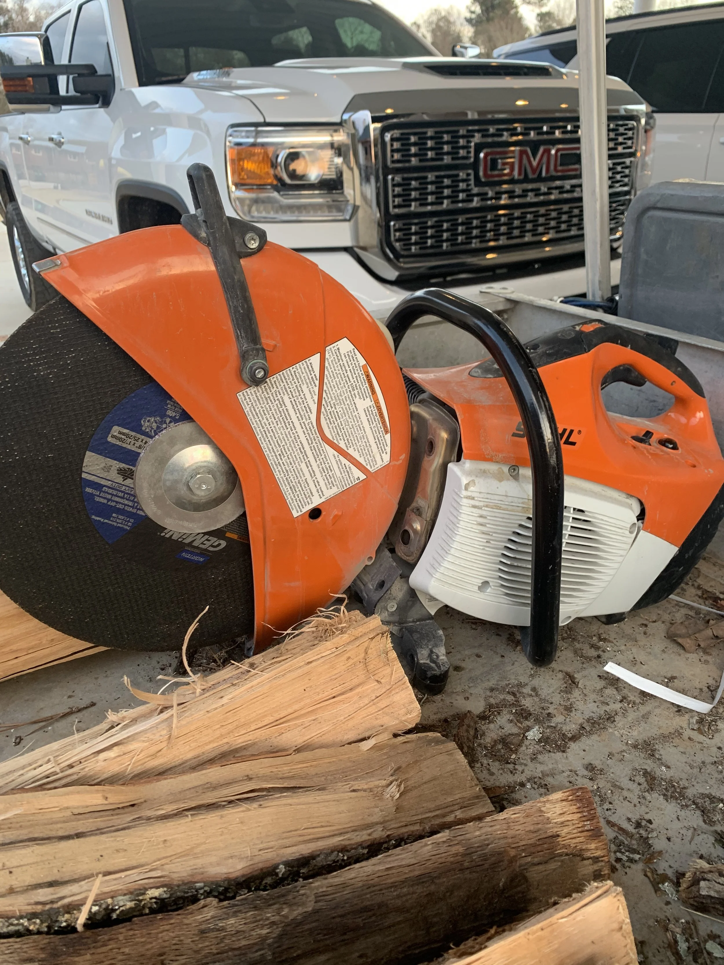 A chainsaw cutting through logs with a GMC truck parked in the background.