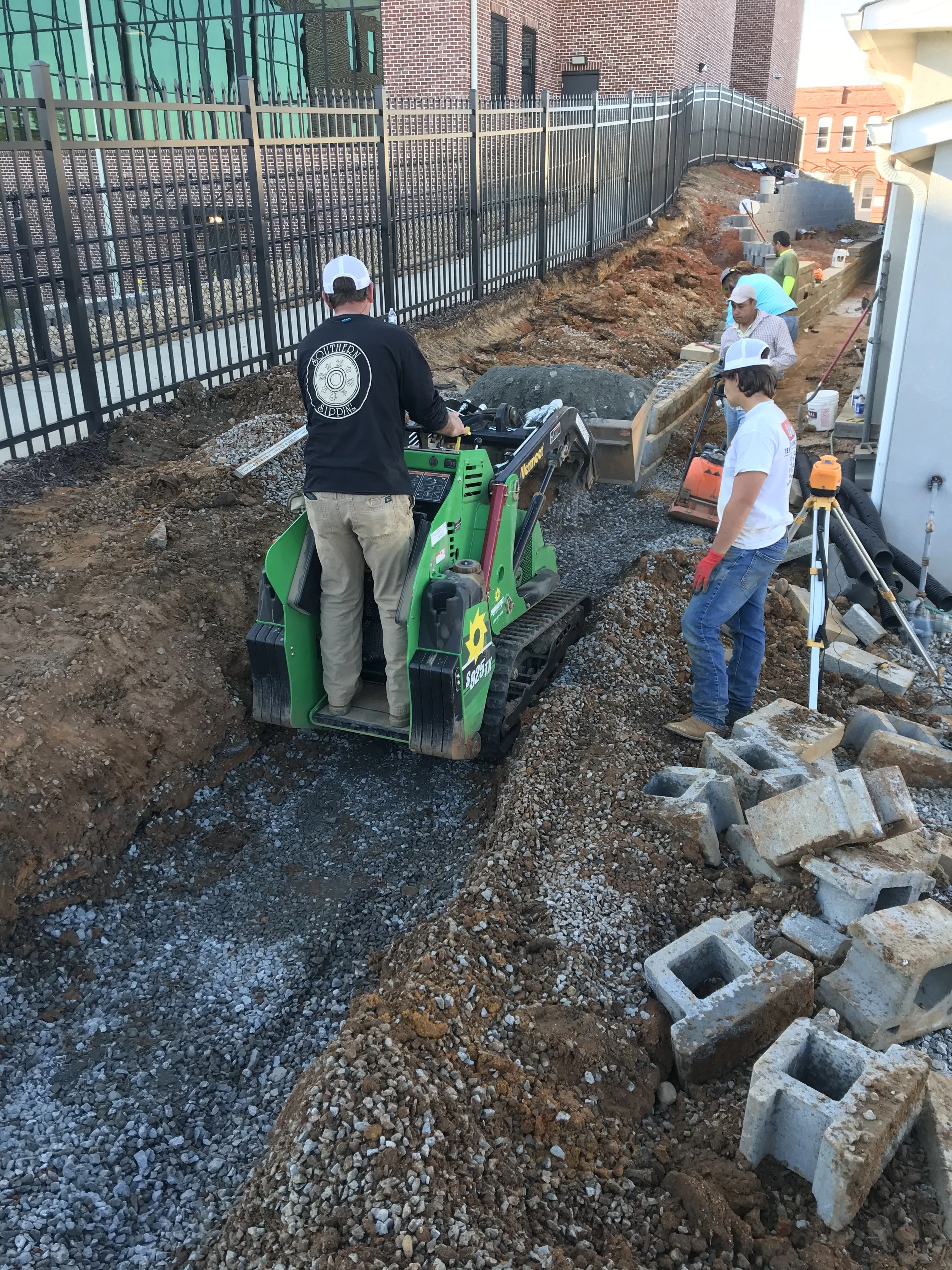 Construction workers and machinery working on a dirt and gravel pathway along a brick building and iron fence, with construction tools and concrete blocks nearby.