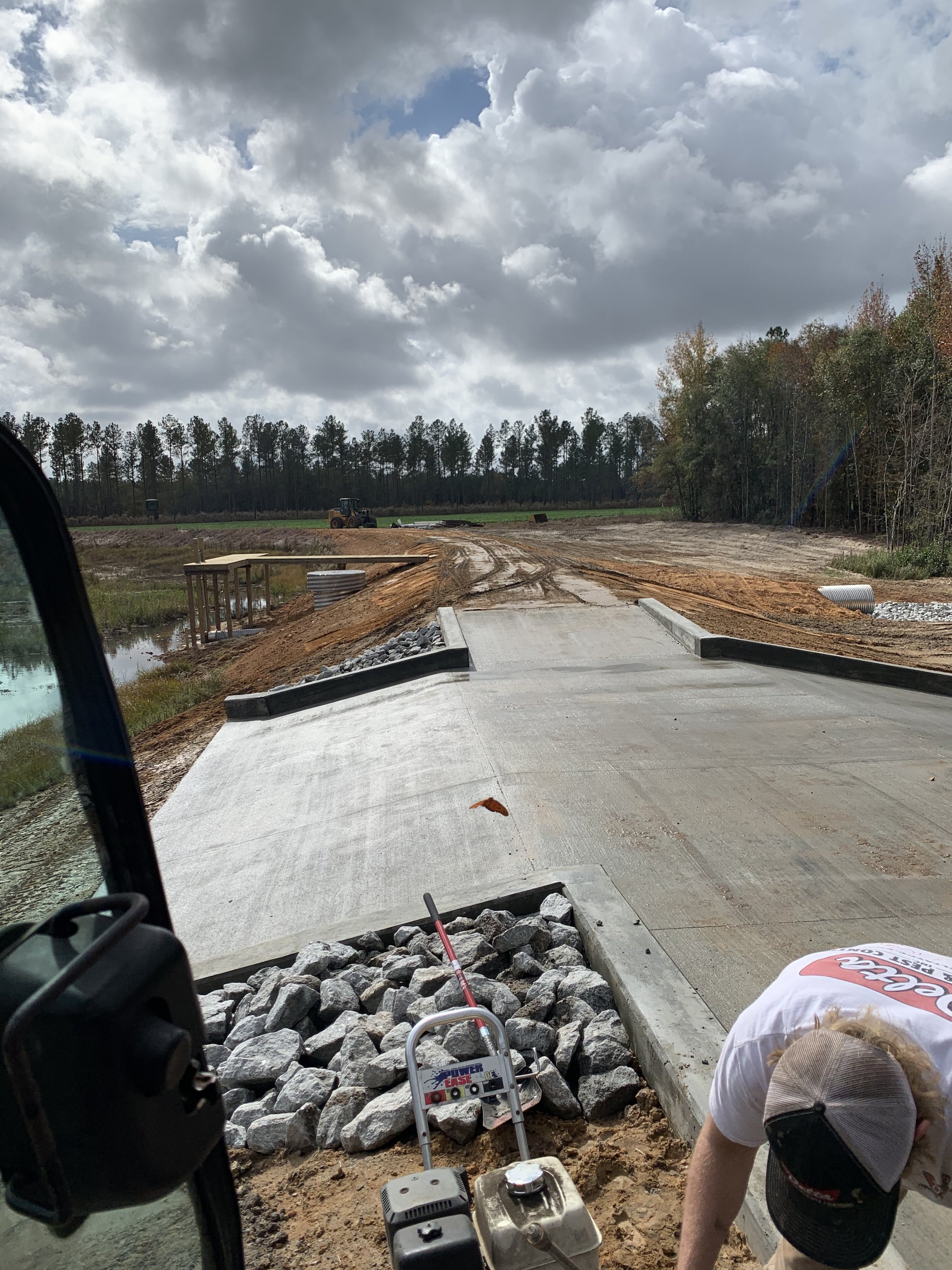 Construction worker working on a newly poured concrete bridge on a rural road, with construction equipment and dirt surrounding the area, under cloudy skies.