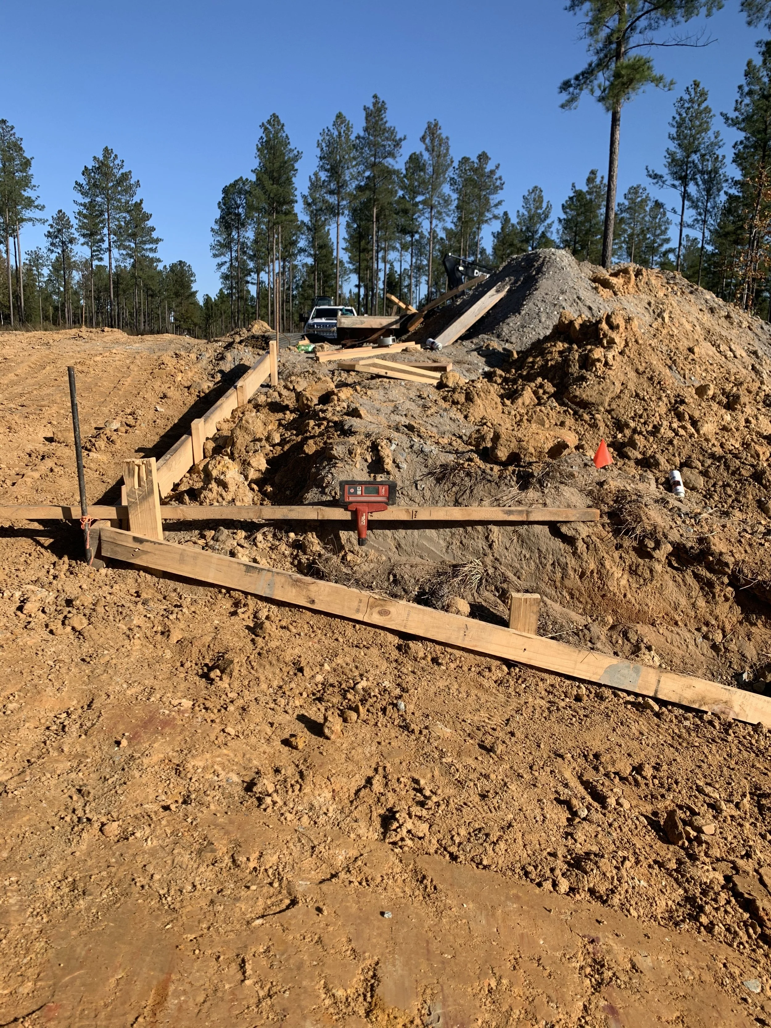 Construction site with wooden formwork set up for a foundation, dirt and soil mounds in the background, and trees under a clear blue sky.