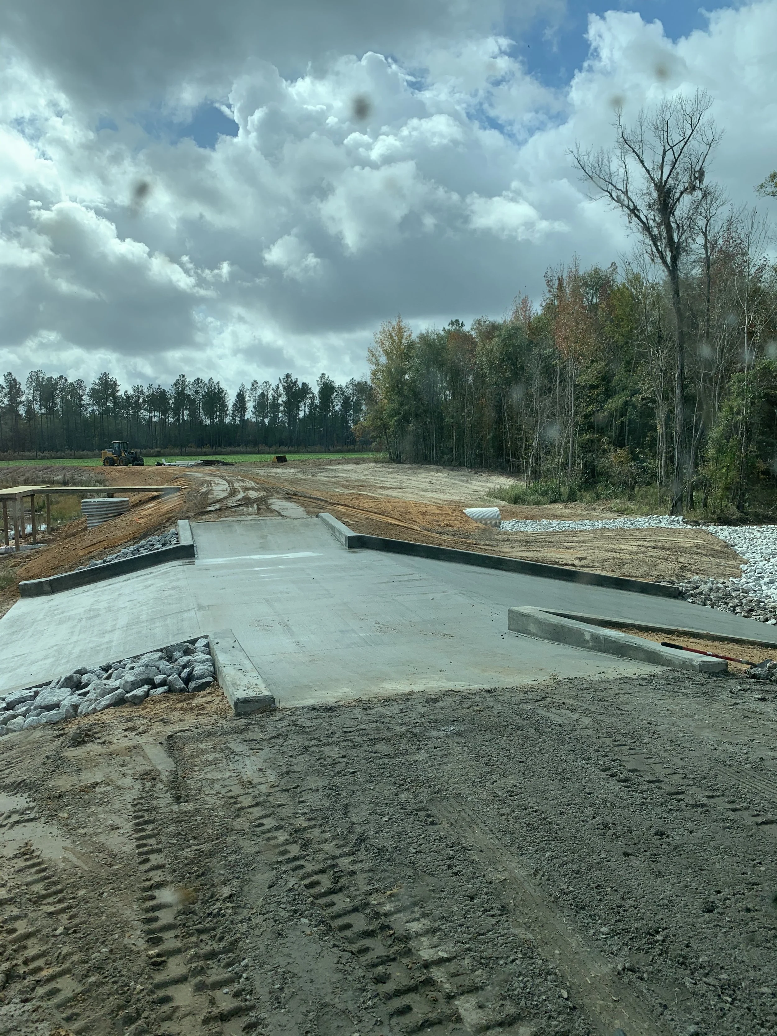 Construction site with a partially built concrete road, dirt and gravel around, a tractor in the background, and cloudy sky overhead.