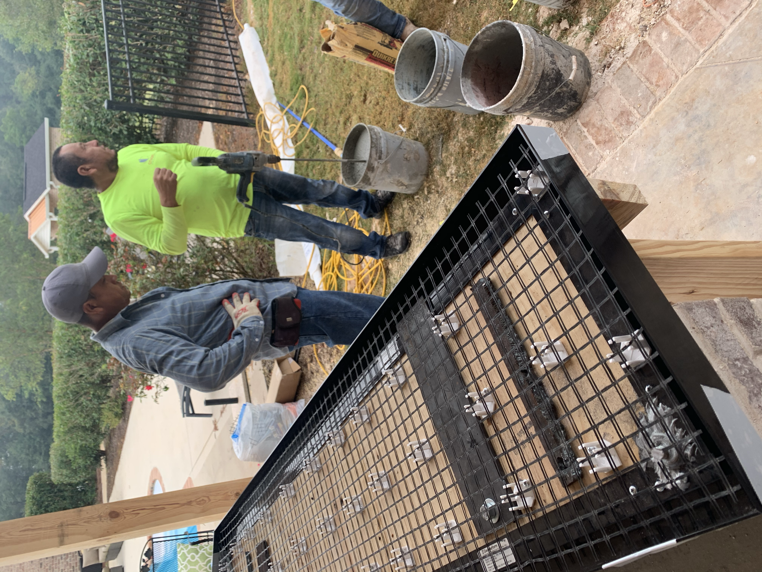 Two men standing at a backyard construction site, one in a neon green shirt and the other in a gray shirt and cap, discussing a construction project. There are buckets, tools, and construction materials nearby, with a grill or outdoor oven in the for