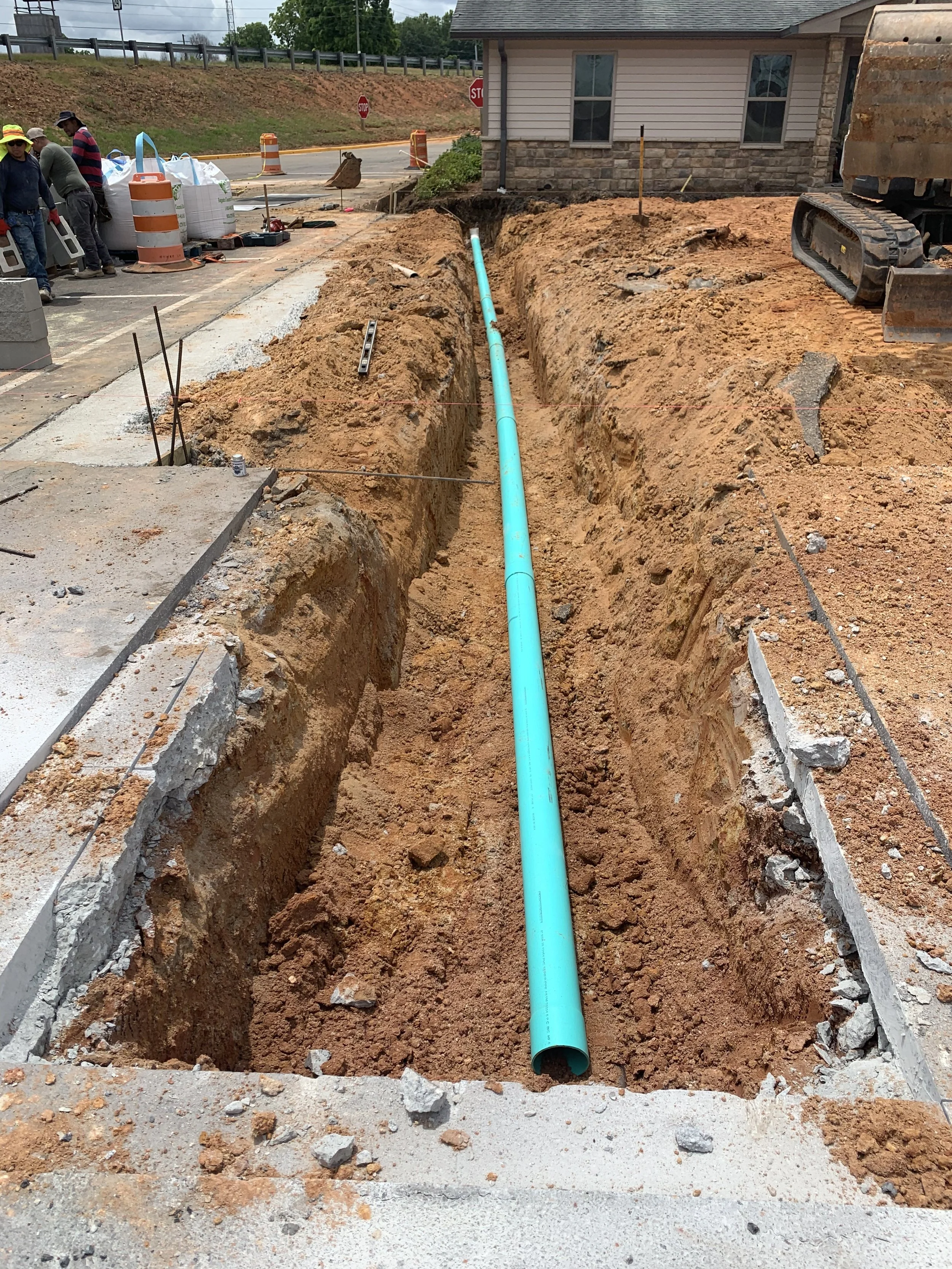 Construction site with a trench for underground piping, a blue pipe running through the trench, and construction workers in the background, along with construction cones and equipment.