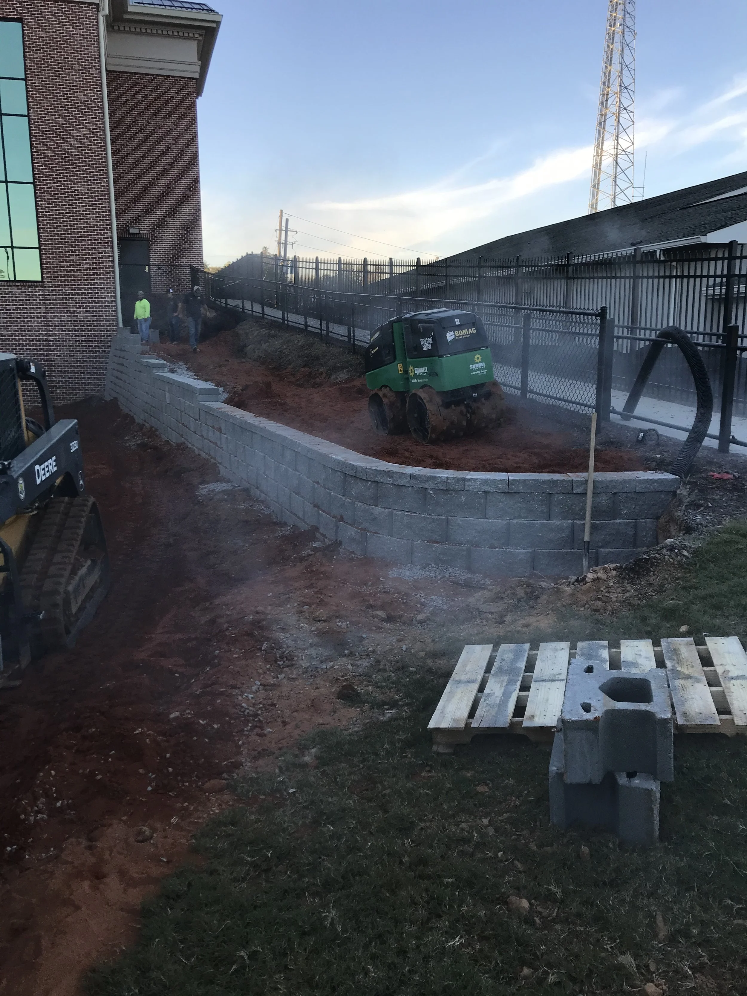 Construction workers building a curved retaining wall with concrete blocks along a sidewalk outside a brick building. There is a small green compact roller used for compacting soil or gravel, and a mini excavator with a bucket attachment. The area is