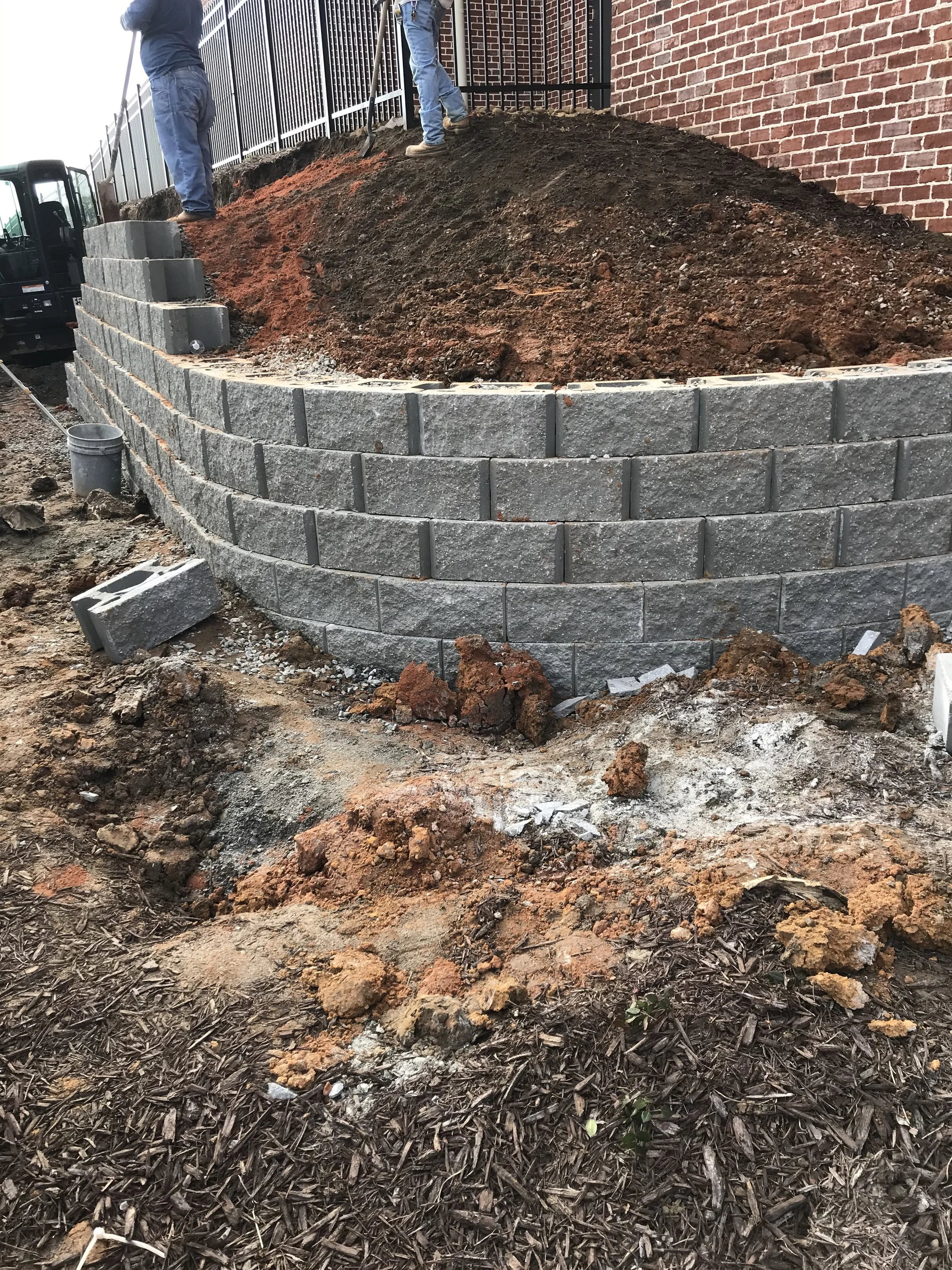 A construction site with a newly built stone retaining wall. Two workers are atop the dirt slope, working near a metal fence attached to a brick building. Construction equipment is visible on the left side of the image.