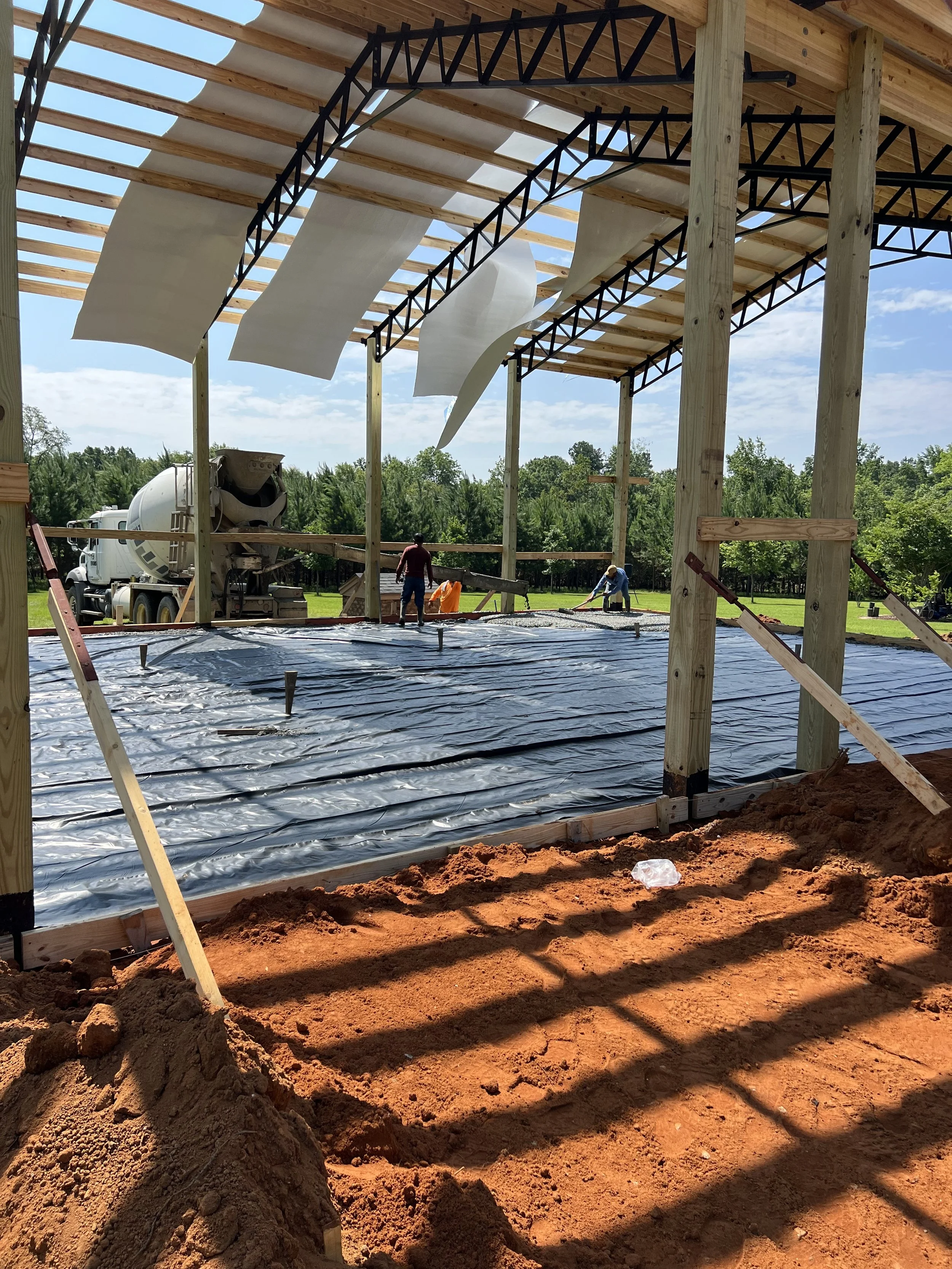 Construction site with a porch being built, workers pouring concrete, and a cement mixer truck in the background. Wooden framework and black roofing underlayment visible.