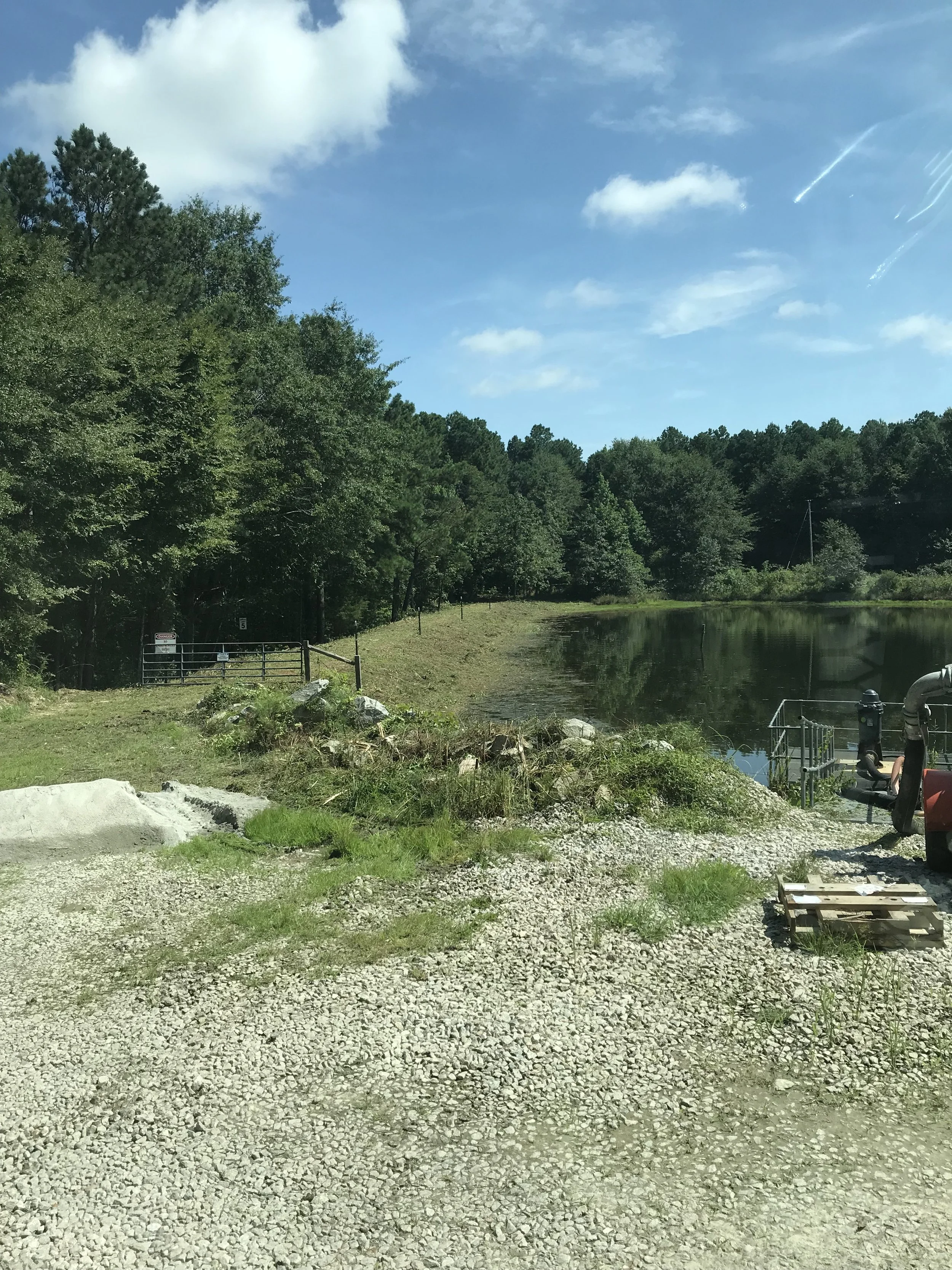 A gravel pathway beside a small body of water, surrounded by green trees, with a partly cloudy blue sky overhead.