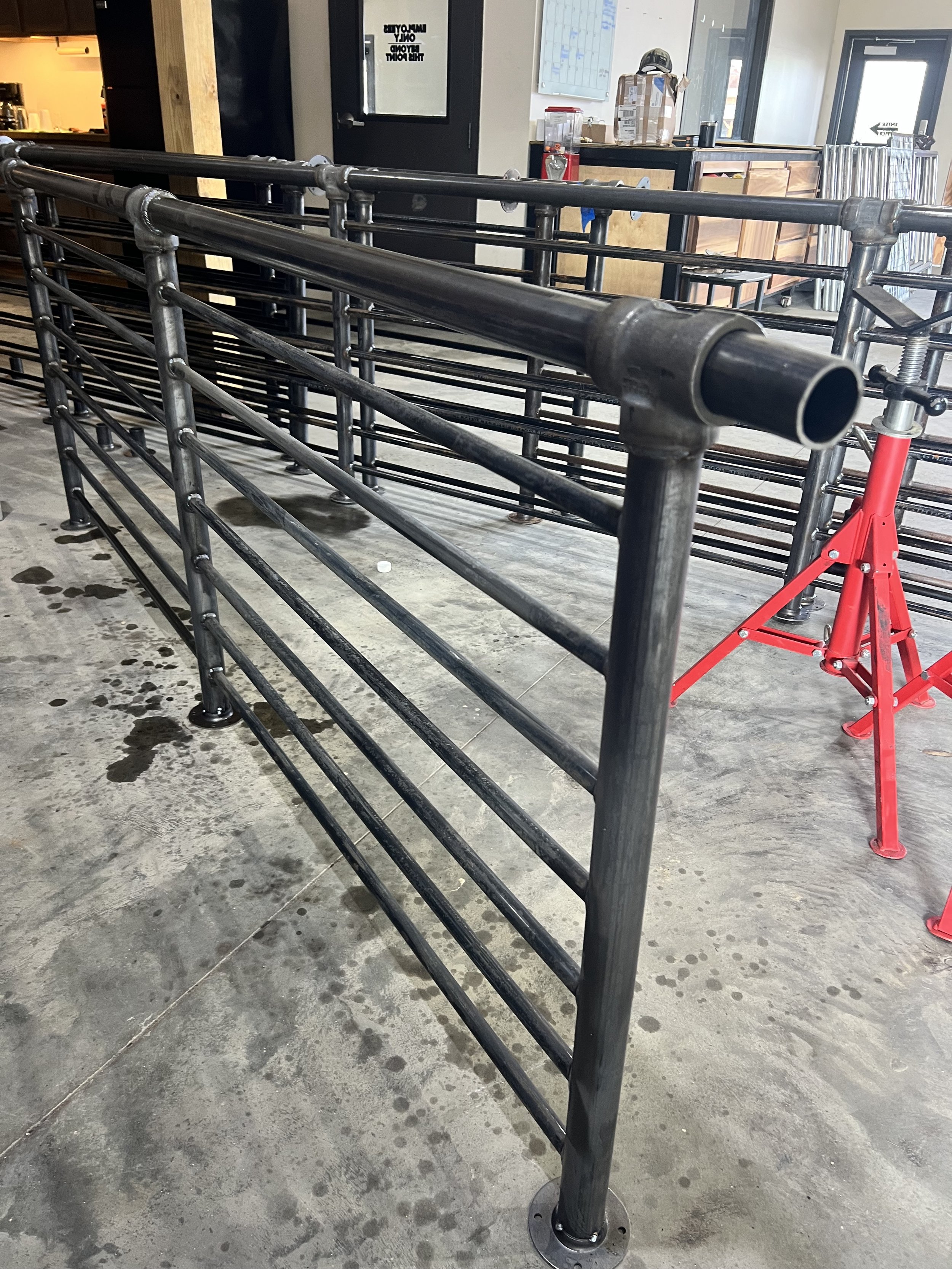 Empty black metal pipe fencing on concrete floor inside a building, with some water spots, and a red jack stand nearby. In the background, there is a counter with various items and a door with signs.