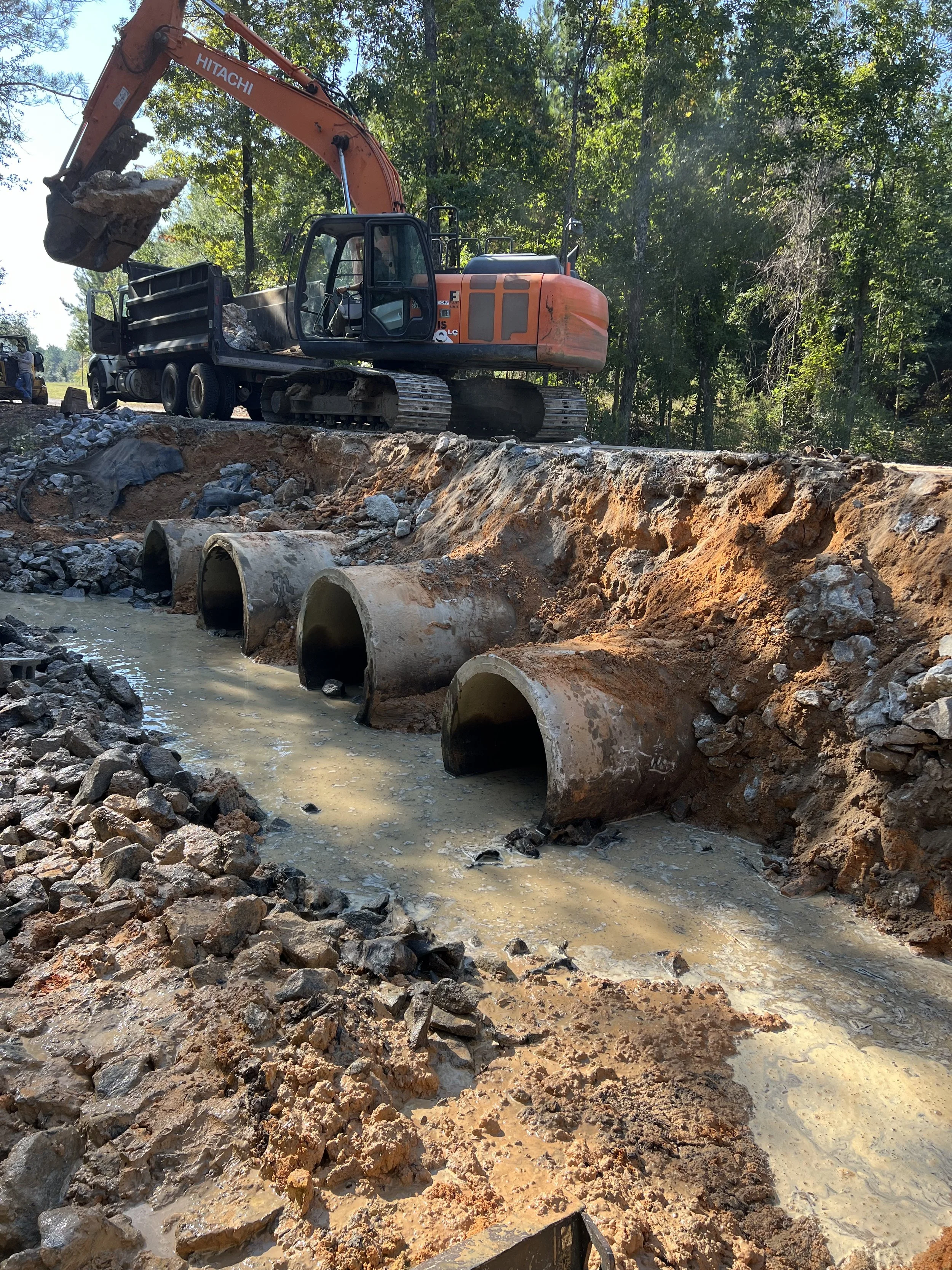 Construction site with large excavator digging a trench and installing large concrete pipes for a drainage or sewer system, surrounded by dirt, rocks, and construction debris, with trees in the background.