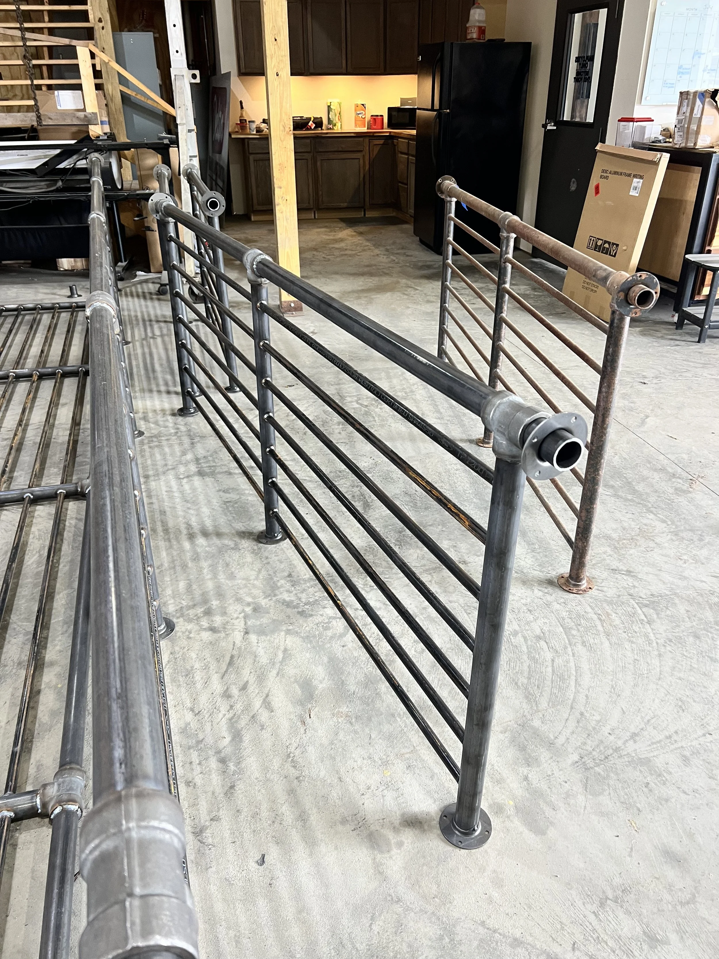 Various metal bed frames and rails are arranged on a concrete floor in an indoor space, possibly a workshop or storage area, with a kitchen and equipment visible in the background.