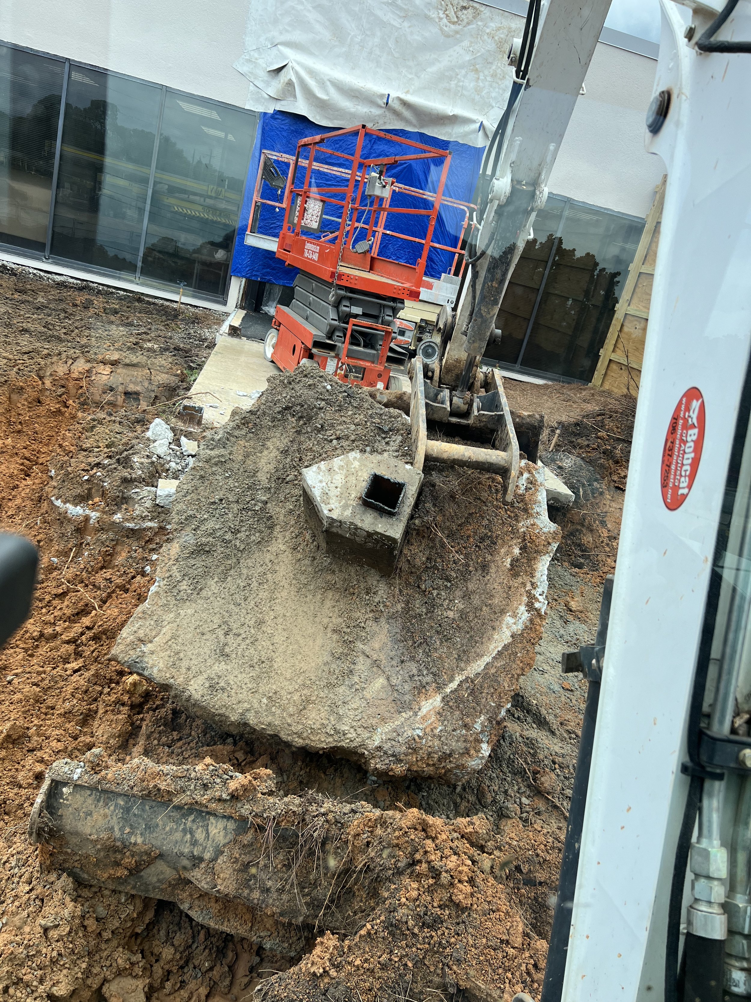 Construction site with an excavator lifting a large, rectangular concrete block. The background shows a building with large glass windows, a blue tarp, and wooden framing.
