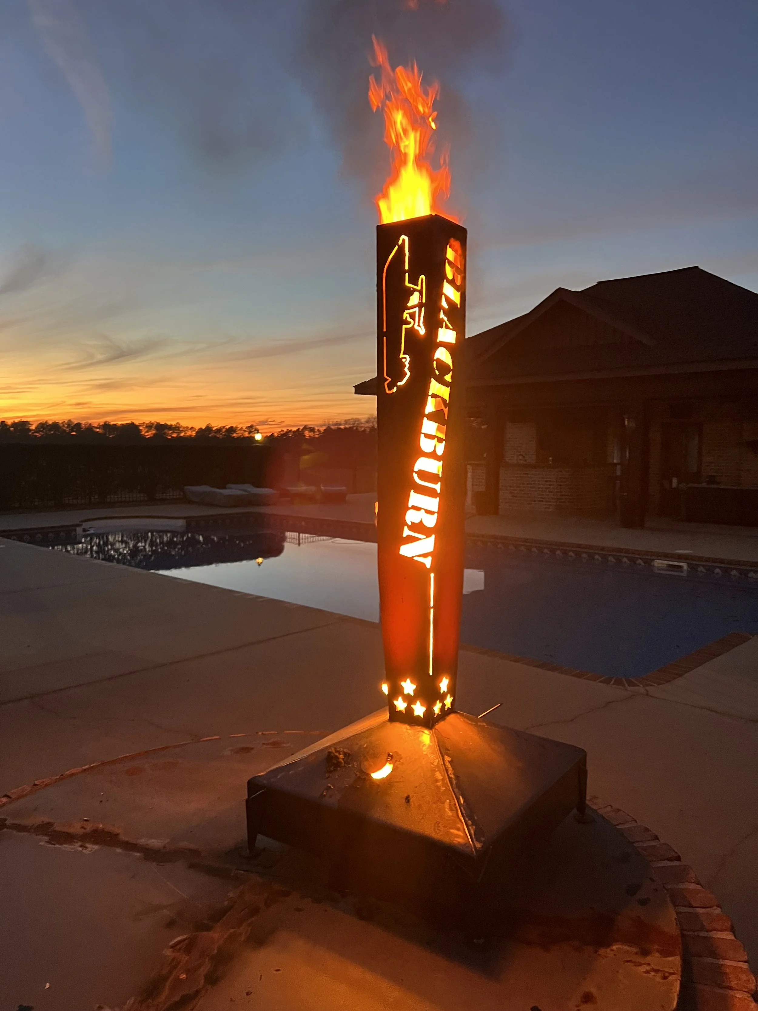 Lit decorative torch with the word 'FOURTH OF JULY' and a flame on top, near a swimming pool during sunset.