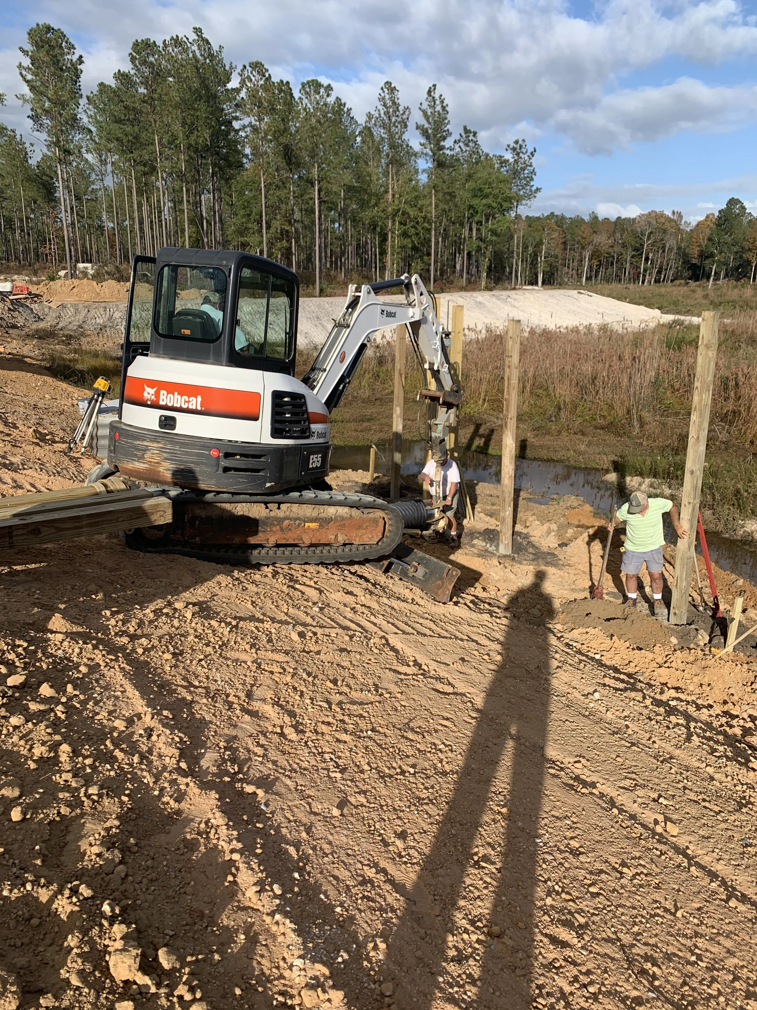 Construction site with a Bobcat mini excavator and two workers installing support beams along a small waterway, dirt and trees in the background.