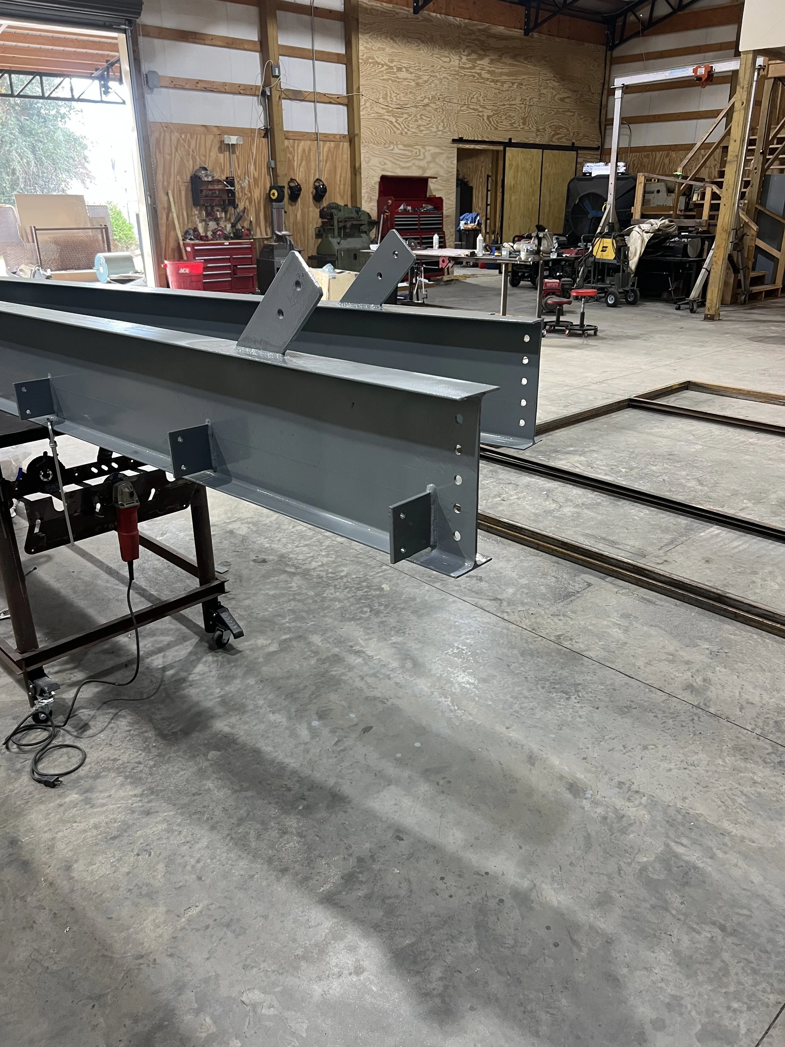 Inside a workshop with metal beams in the foreground and various tools and equipment on workbenches and storage cabinets in the background.
