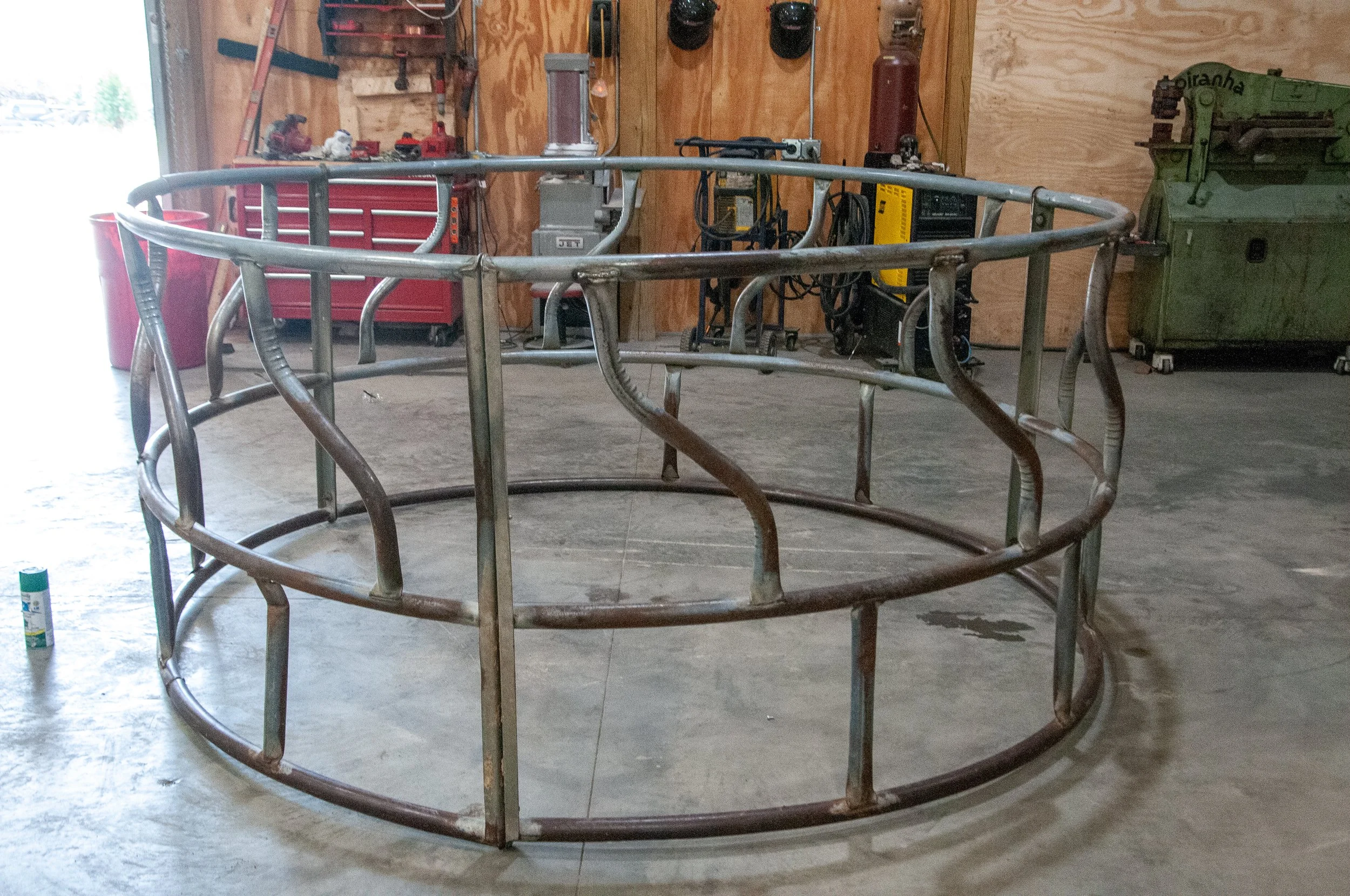 A large circular metal structure, possibly a frame, is placed on the concrete floor inside a workshop or garage. The background shows various tools and equipment including a red tool chest, a green machine, and a welding machine, with plywood walls.
