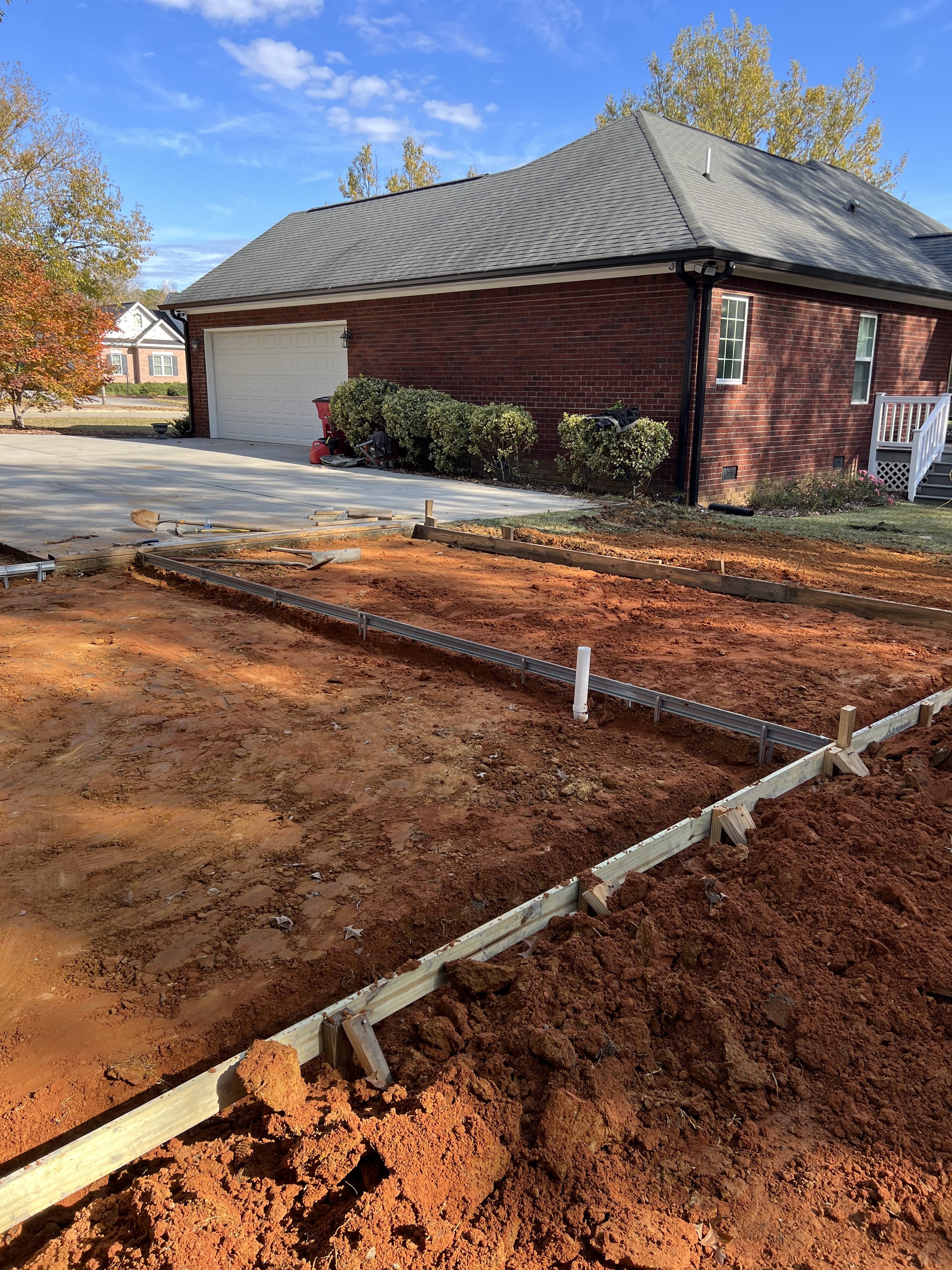Construction site in front of a suburban house with a brick exterior, next to a driveway, and framed for a new patio or foundation. The sky is partly cloudy with trees showing fall foliage.