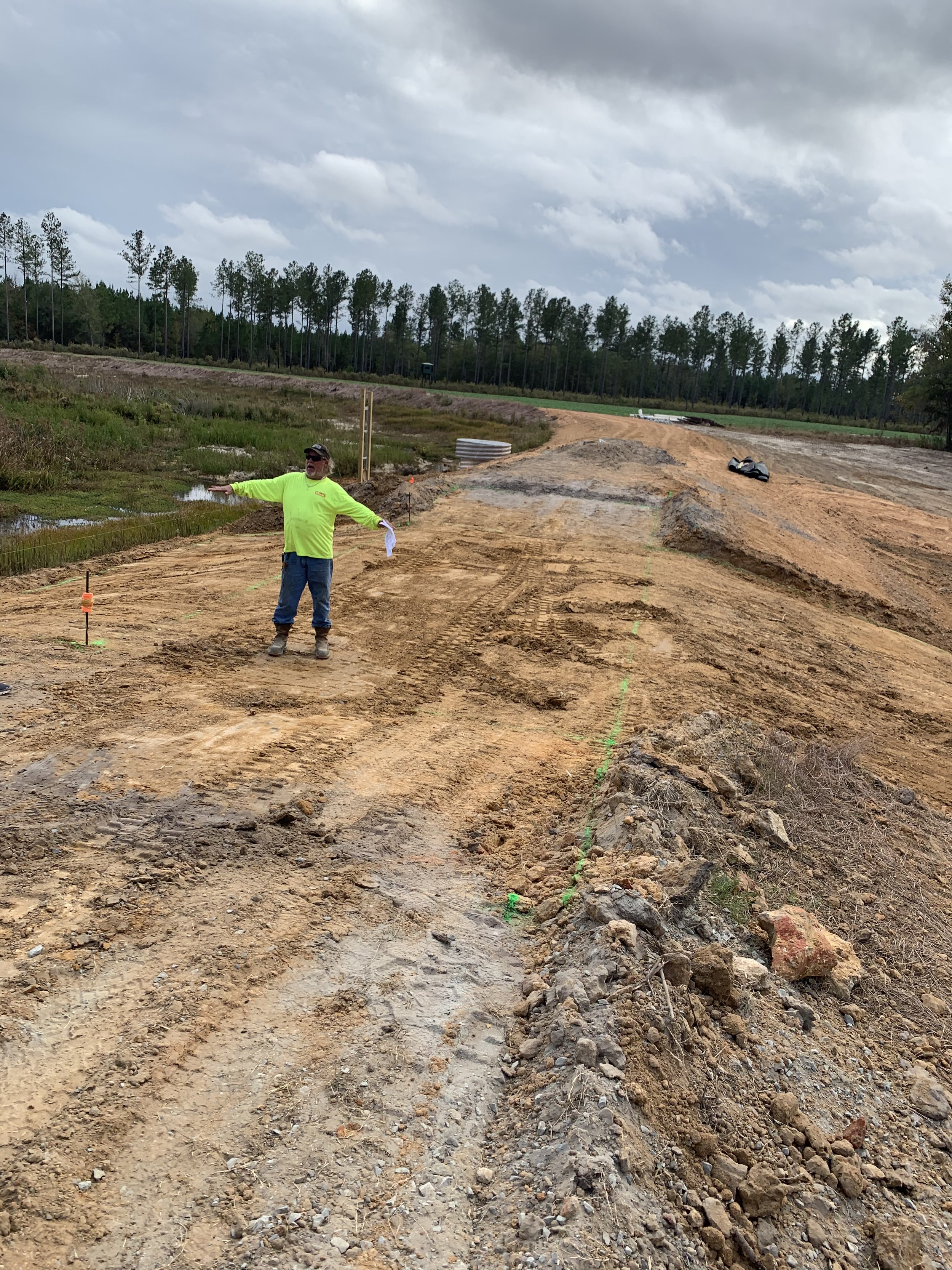 A construction worker in a bright yellow-green shirt and safety glasses stands on a dirt road under an overcast sky. The worker is holding a piece of paper and pointing towards the distance, with construction markings on the ground nearby.