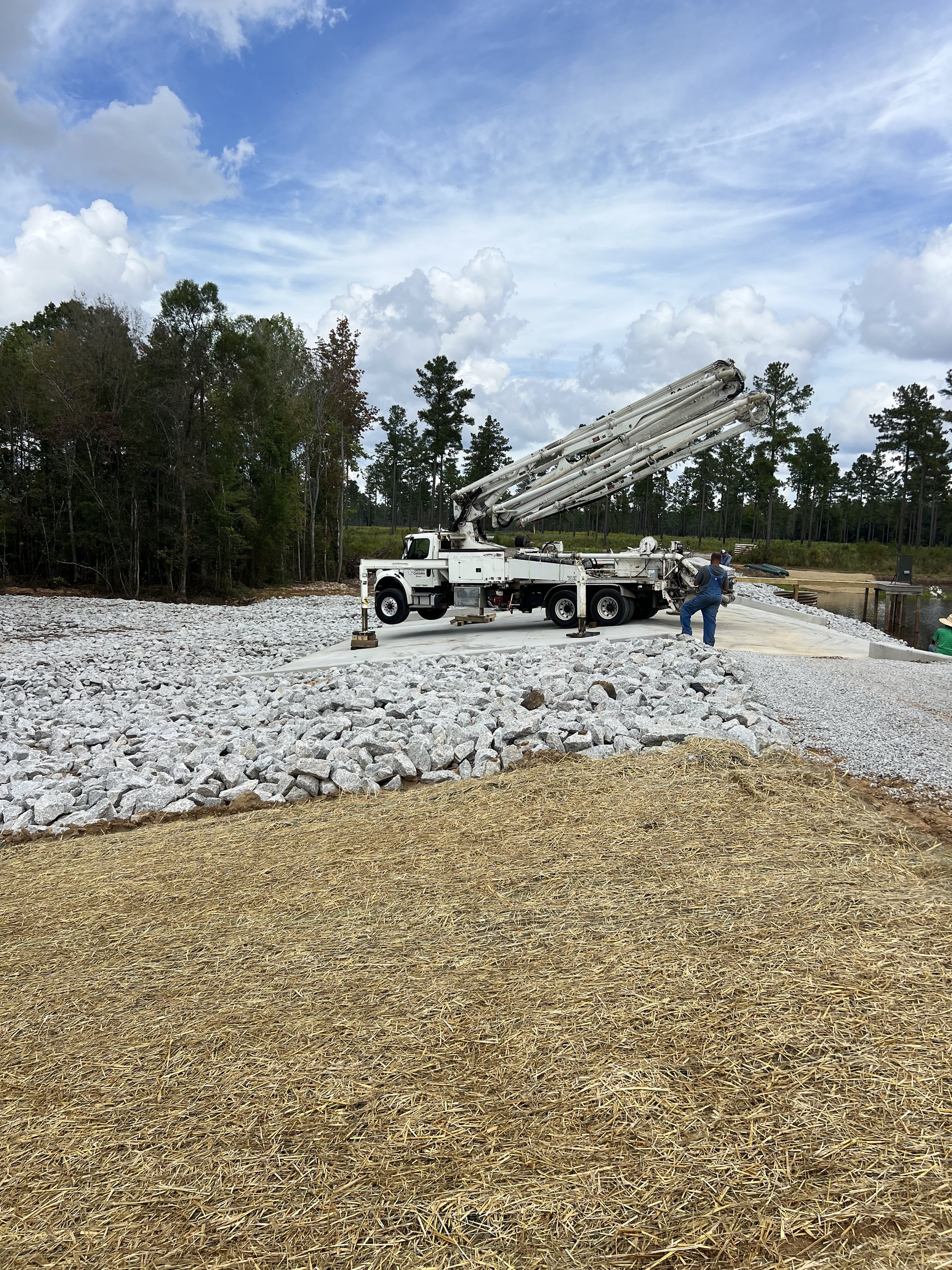 Construction site with a concrete truck and workers laying rocks and gravel for groundwork, set outdoors with cloudy sky and trees in the background.