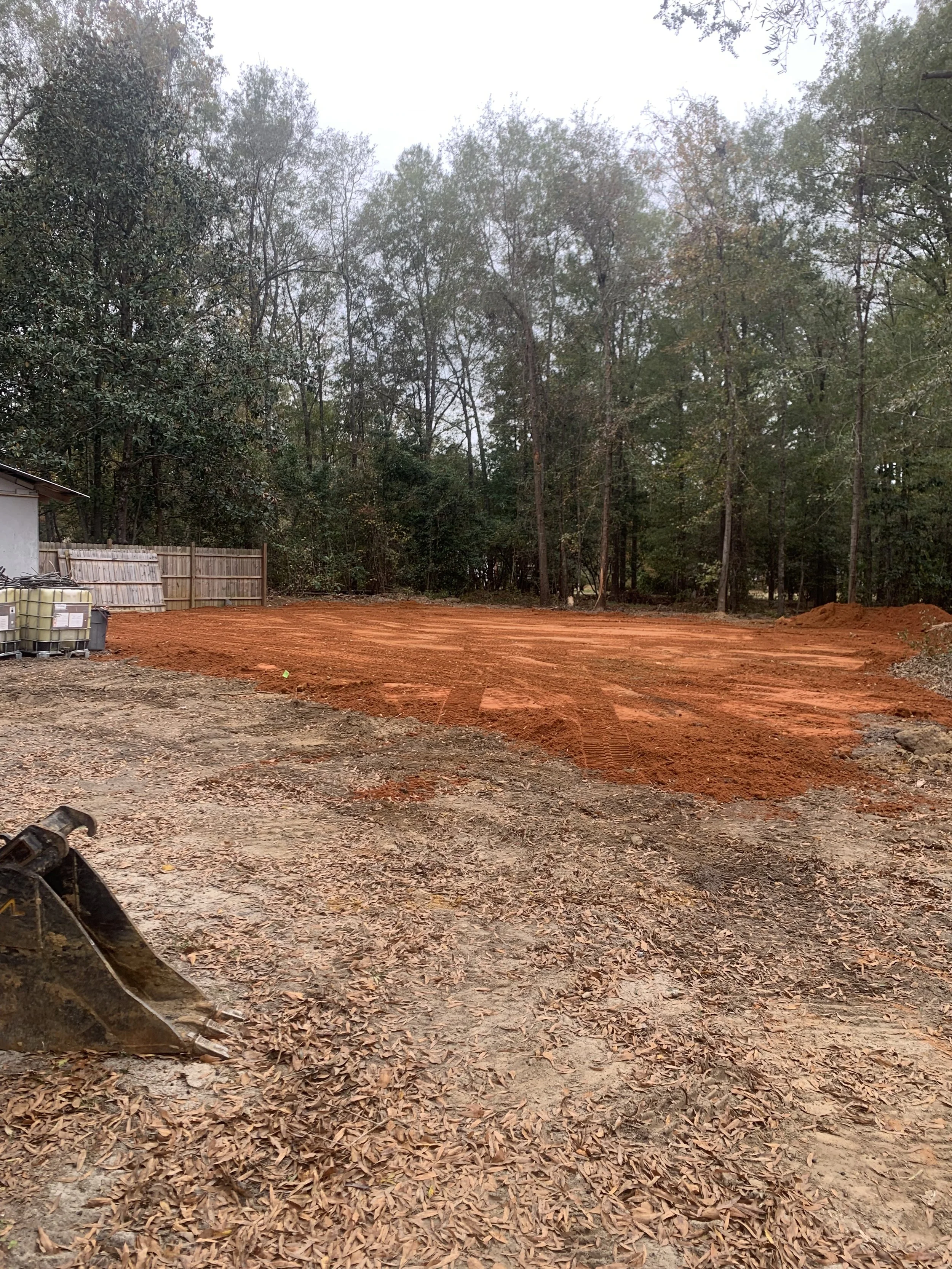 A construction site with red dirt, some construction equipment, and a background of trees and a wooden fence.