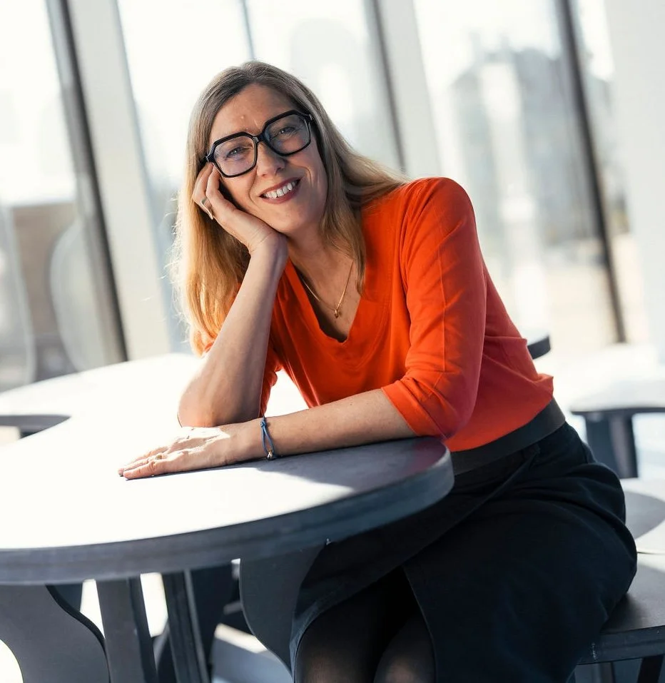 Eine lachende Frau mit Brille sitzt an einem runden Tisch in einem modernen Bürogebäude, hinter großen Fenstern.