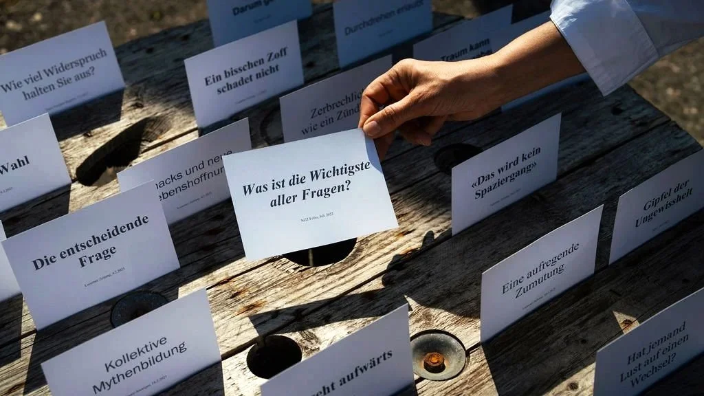 Hand, die eine Karte mit der Frage „Was ist die Wichtigste aller Fragen?“ auf einem Holztisch hält, umgeben von weiteren Karten mit Fragen wie „Die entscheidende Frage“ und „Kollektive Mythenerbildung“.