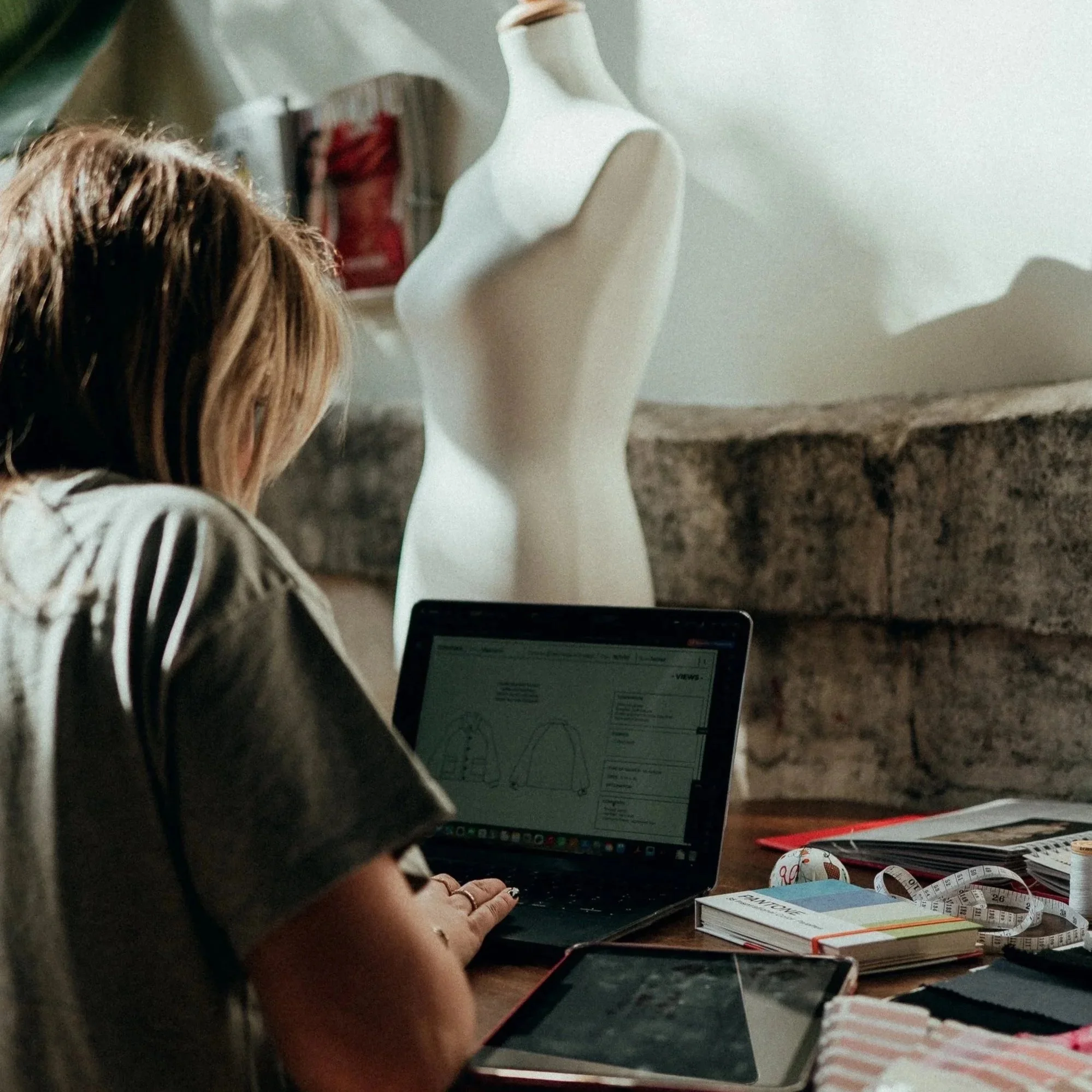 Une femme travaille sur un ordinateur portable avec des schémas de vêtements à côté, dans un environnement intérieur, avec un mannequin en toile dans l'arrière-plan.