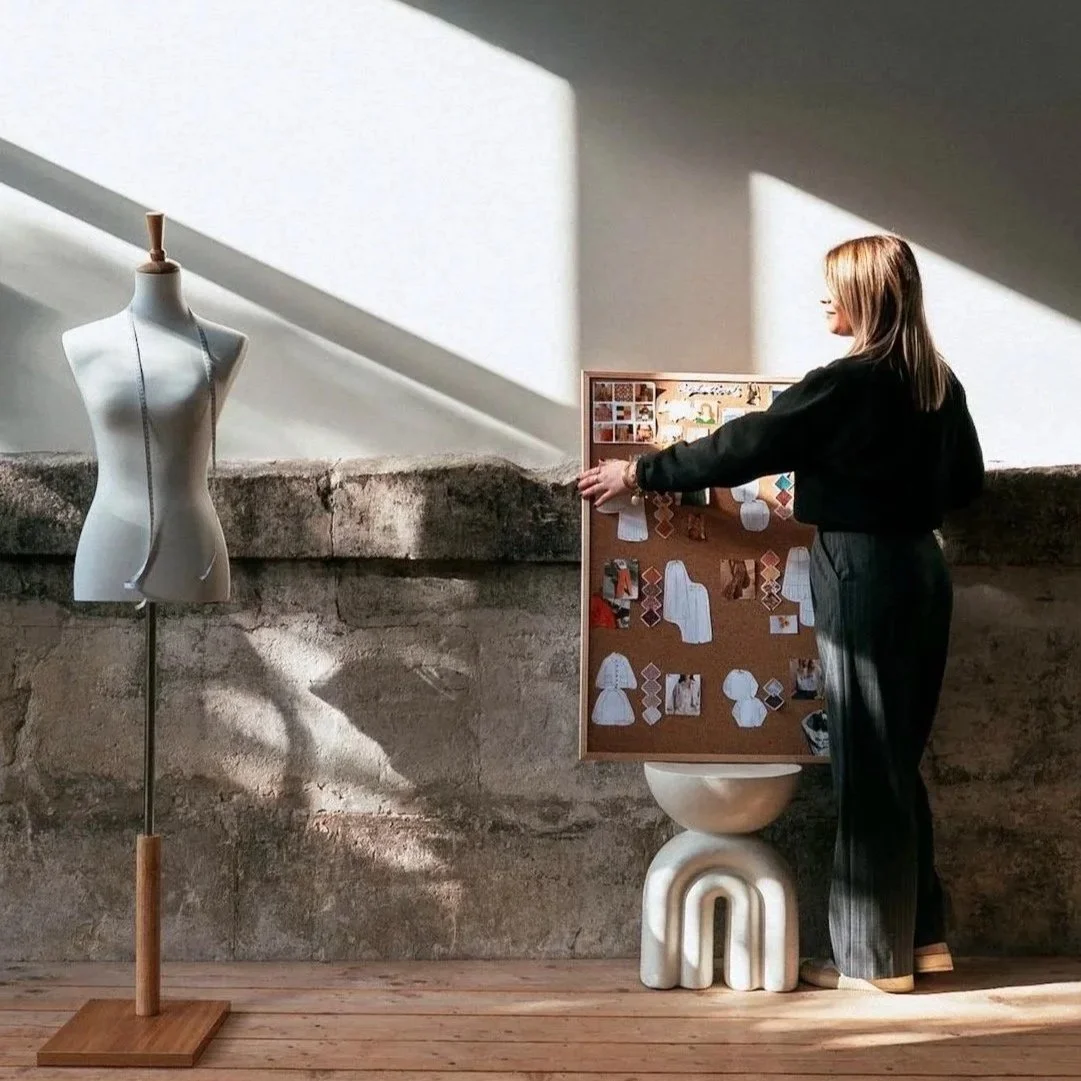 Une femme se tient devant un tableau avec des images de patrons de couture, à côté d'un mannequin en tissu blanc. La scène est éclairée par la lumière naturelle faisant des jeux d'ombres sur le mur en pierre.