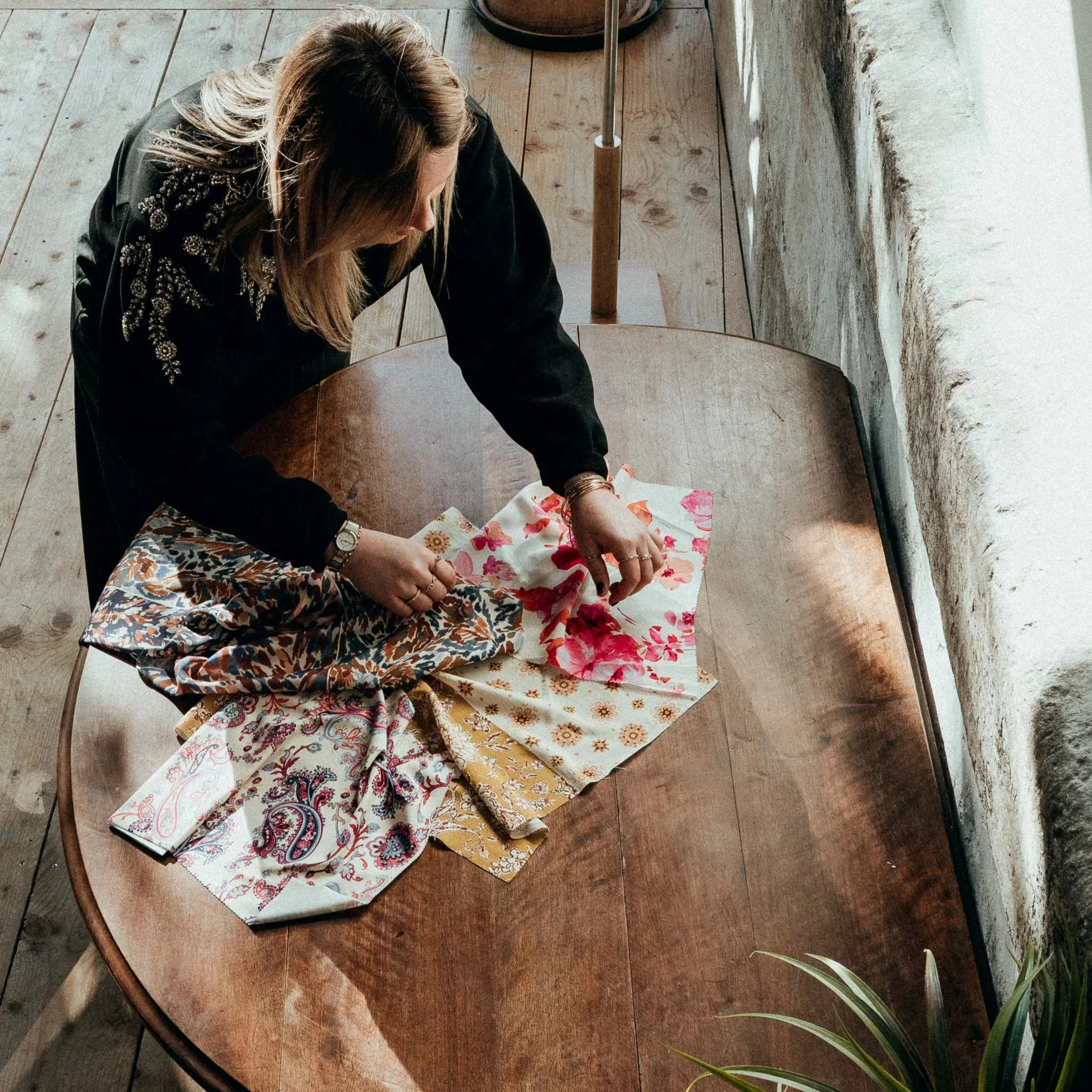 Une femme plie des tissus à motifs floraux sur une table en bois dans une pièce éclairée par la lumière naturelle, près d'un mur blanc en pierre.