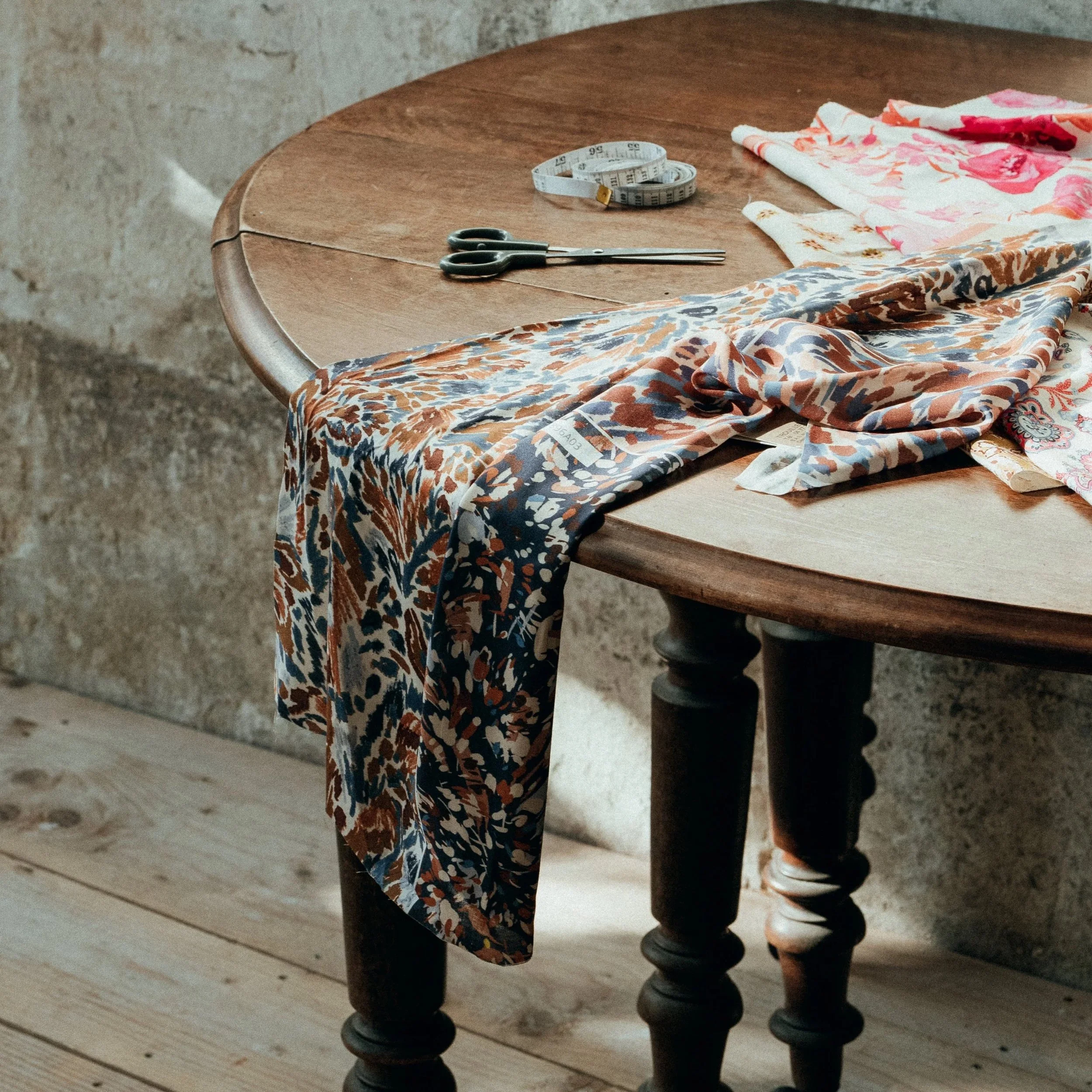 Une table en bois avec un tissu de couture, des ciseaux, un ruban à mesurer, et des morceaux de tissu floral, dans une pièce avec un mur en pierre.