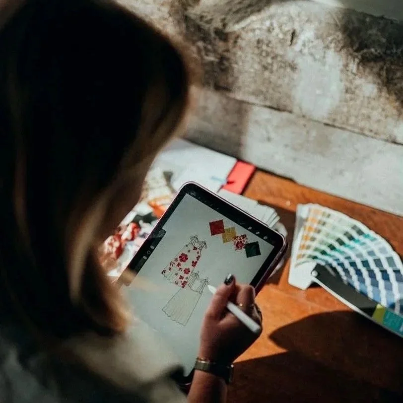 Une femme utilise une tablette pour créer un design de mode avec des échantillons de tissus et une fan de nuancier à côté.