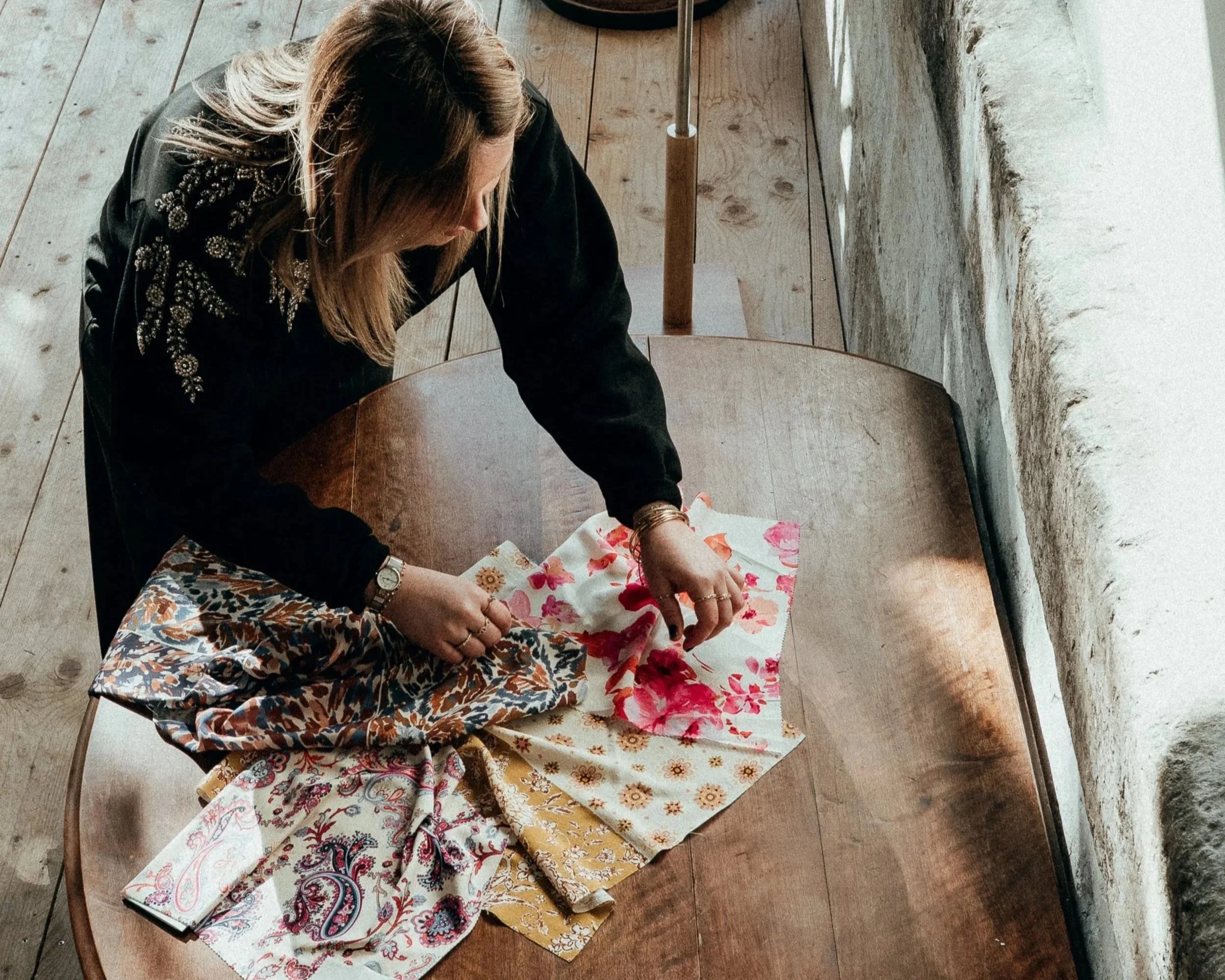 Femme examinant plusieurs tissus colorés avec des motifs floraux et Paisley sur une table en bois.