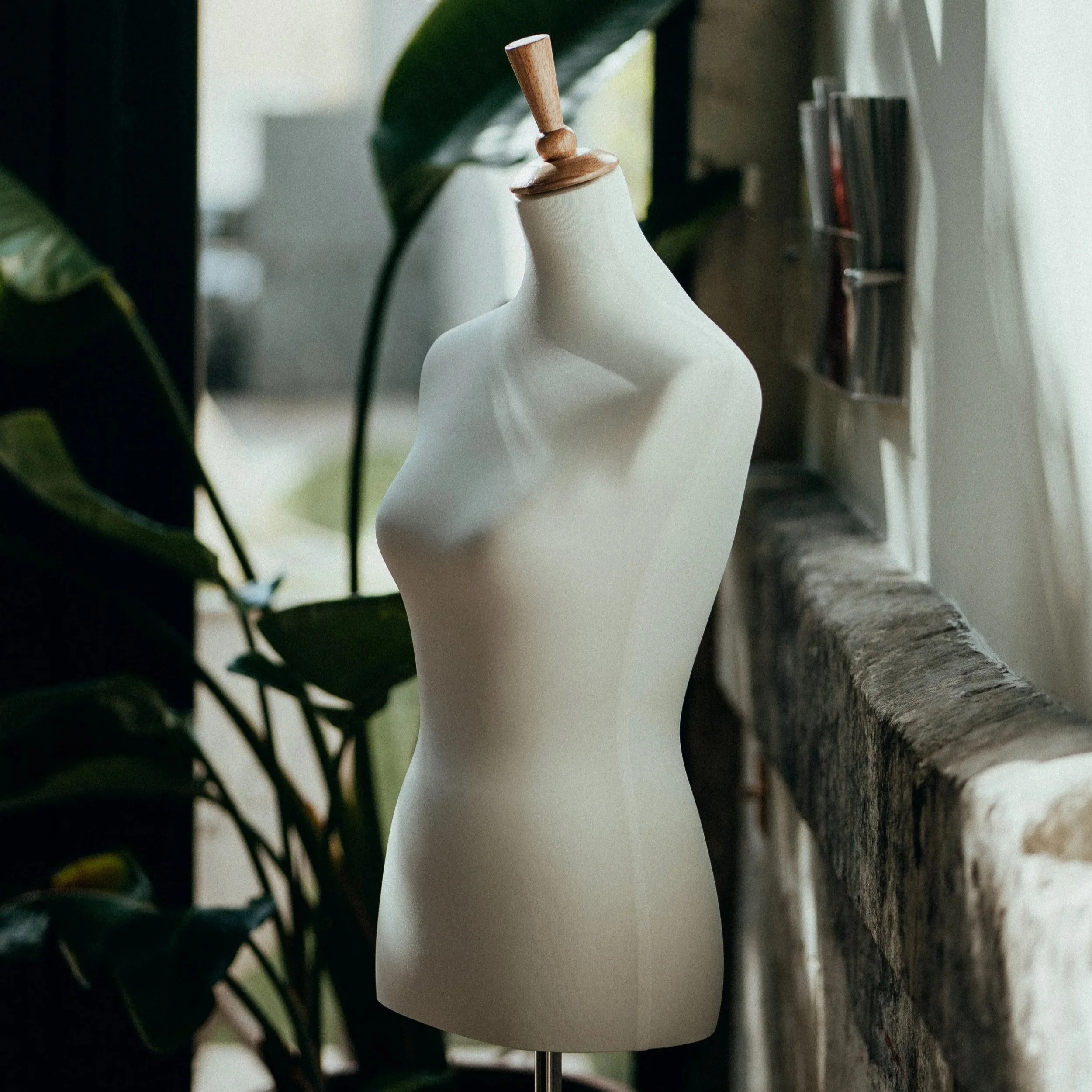 Mannequin féminin blanc sans bras ni tête, placé près d'une fenêtre, avec des plantes vertes en arrière-plan.