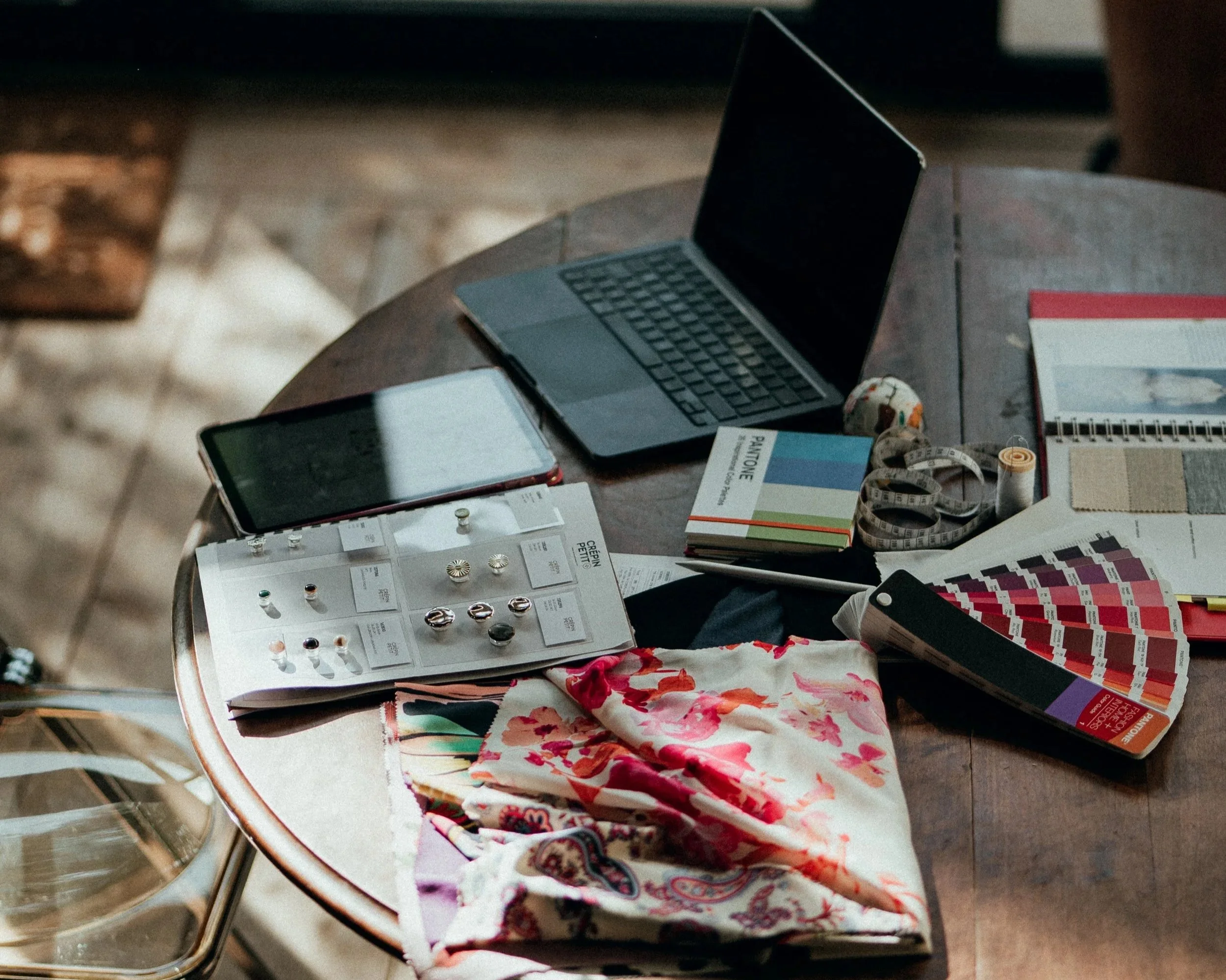 Table en bois avec des échantillons de bijoux, un smartphone, un ordinateur portable, un carnet de peinture, un ruban à mesurer, une palette de couleurs, un échantillon de tissu, et d'autres documents.