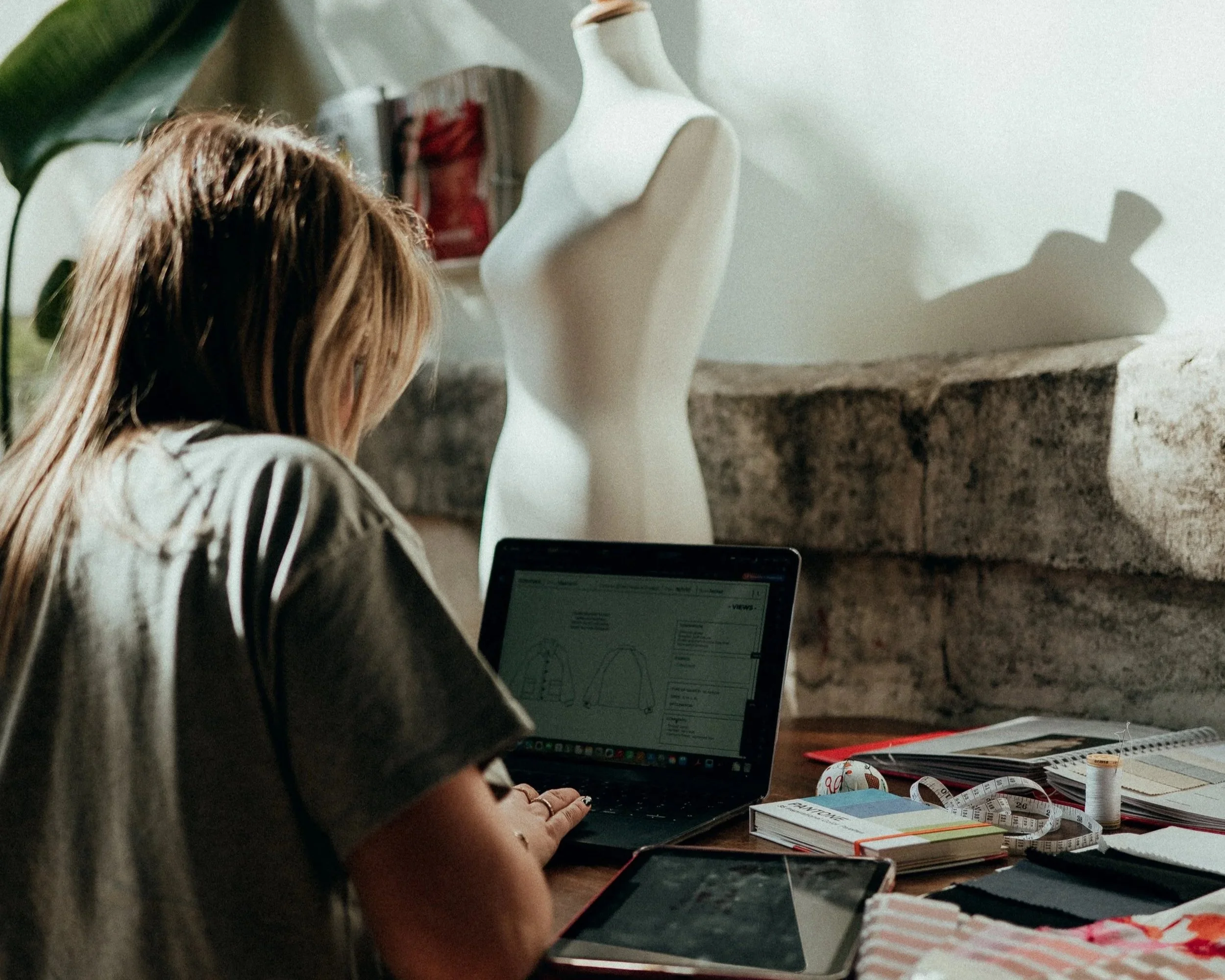 Une femme travaille à un bureau avec un ordinateur portable, des livres, une règle, un ruban à mesurer, une fiche, un smartphone, des bobines de fil, et un mannequin en argile ou en plâtre, dans une pièce éclairée naturellement.