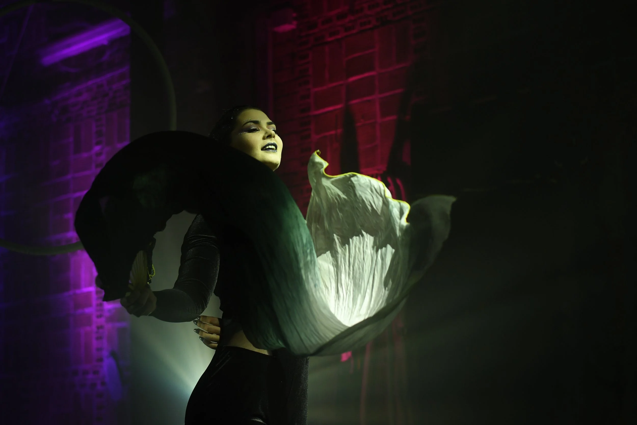 Une femme avec un maquillage sombre porte un costume noir et un grand accessoire blanc qui ressemble à une fleur ou une aile, dans un environnement sombre avec un éclairage coloré violet et vert.