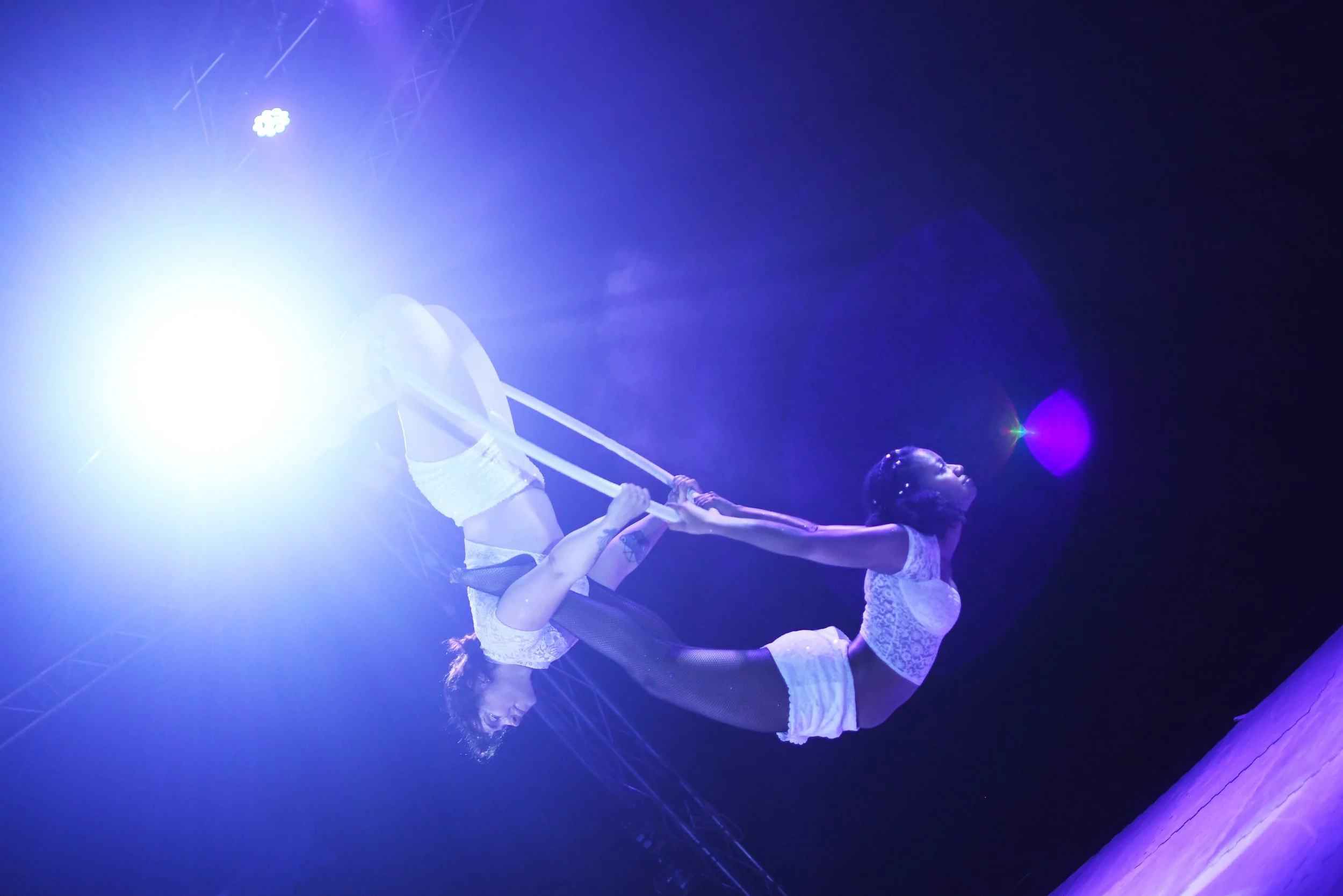 Deux femmes en tenues de danse, debout face à face, en équilibre sur une jambe, tenant un bâton avec des lampes violettes et bleues derrière elles dans un spectacle de danse ou de cirque