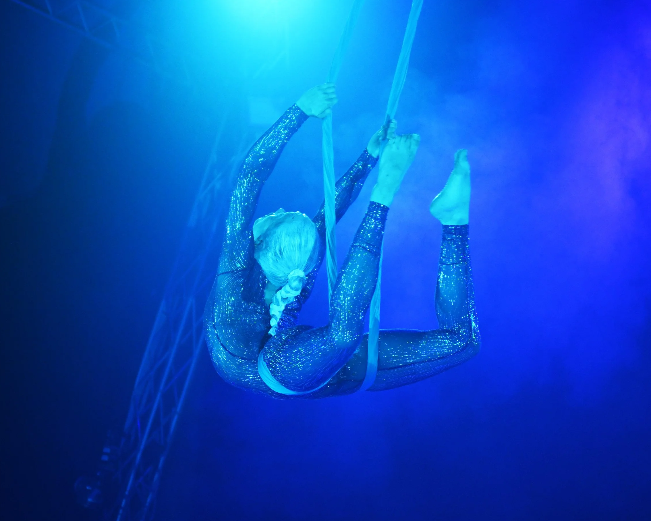 Une performance artistique sous une lumière bleue avec une personne portant un costume scintillant, suspendue par des cordes dans un espace sombre.