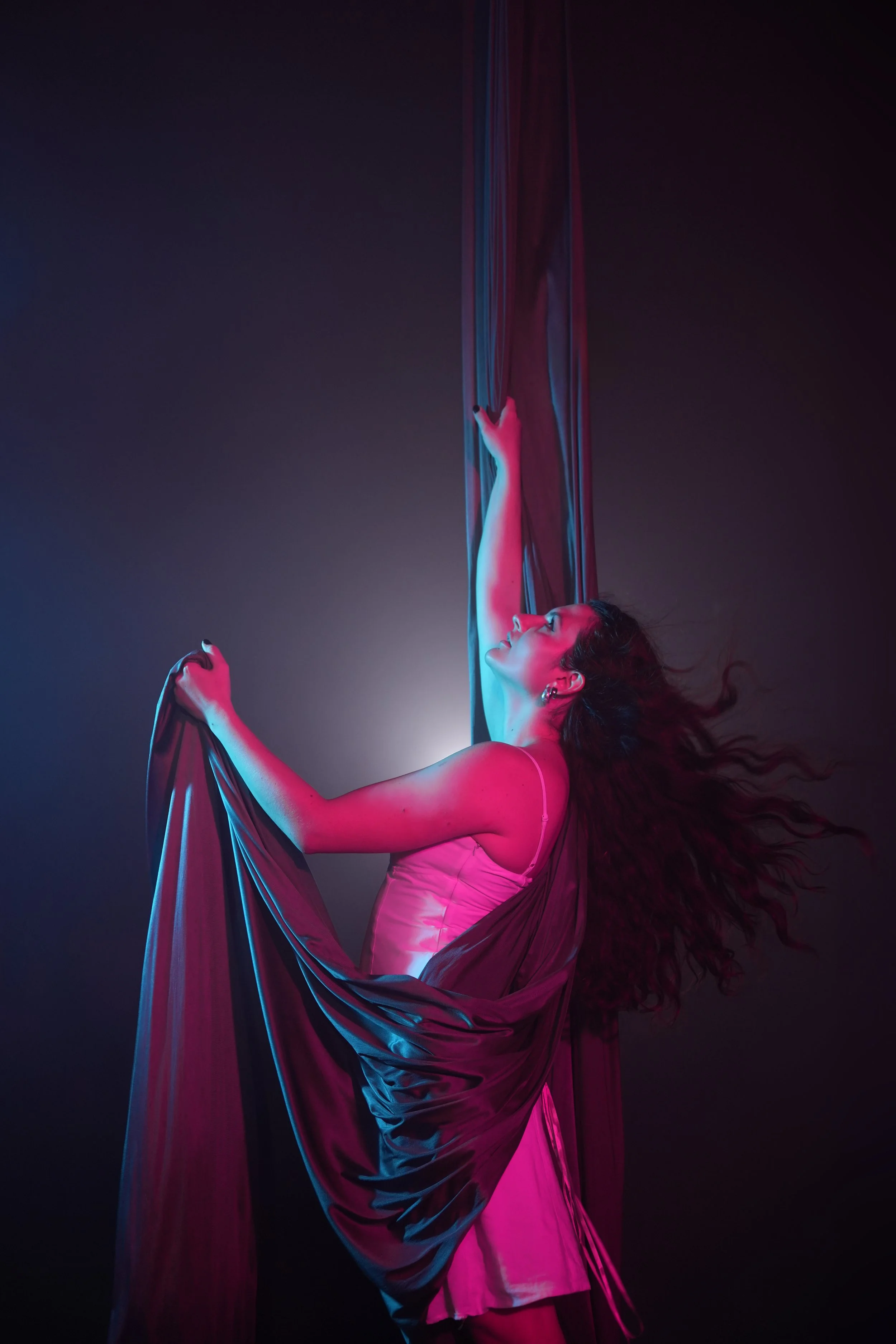 Une femme danseuse suspendue à une toile aérienne dans un éclairage bleu et rose.
