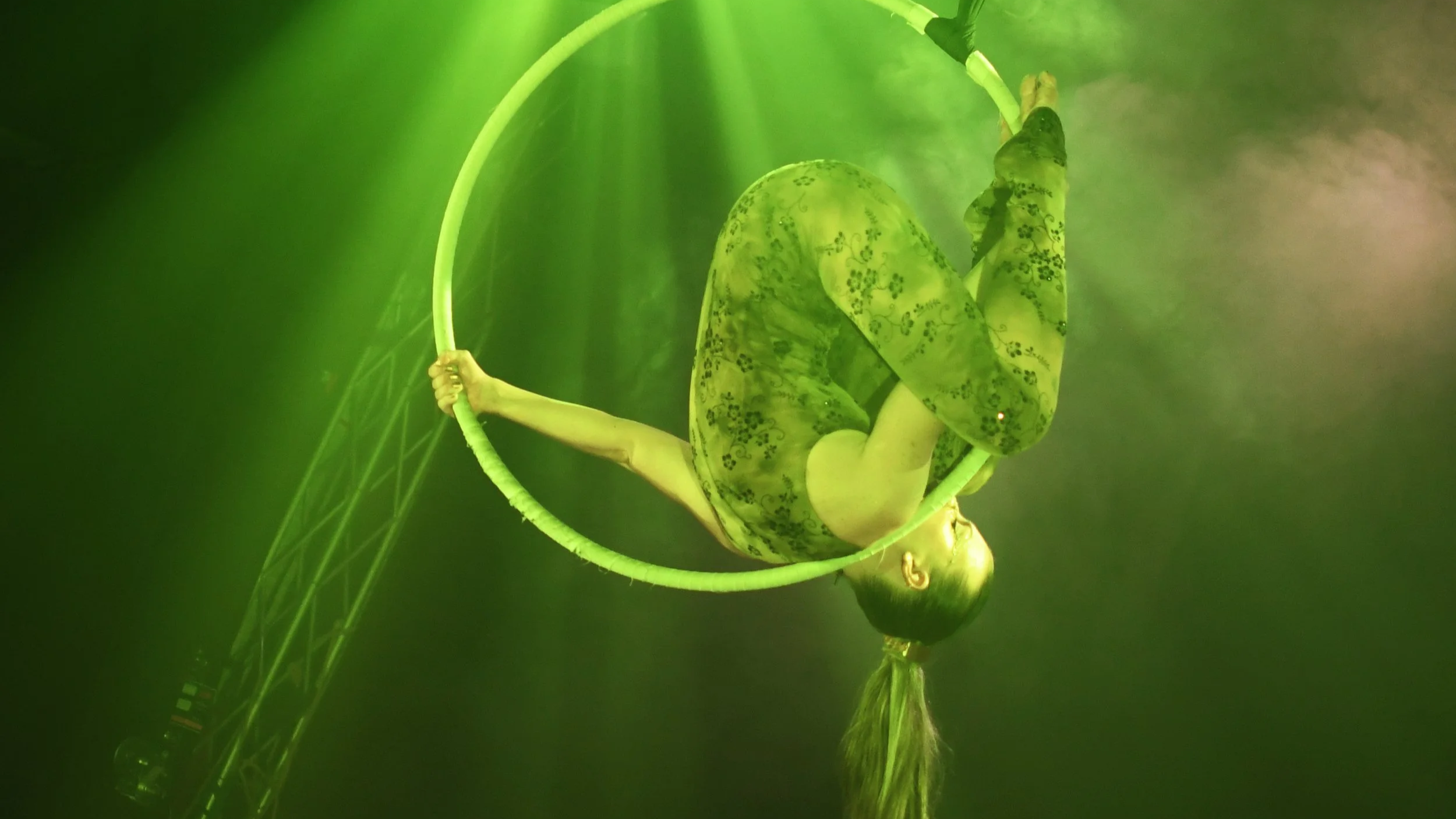 Une acrobate suspendue à un anneau lors d'une performance sous un éclairage vert intense.
